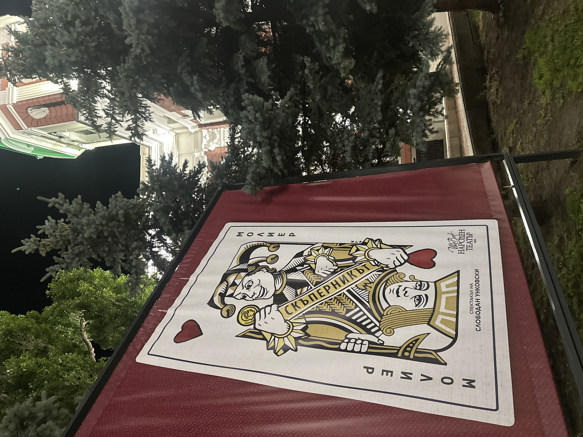 A large banner displaying a playing card, specifically the Jack of Hearts, is positioned outdoors at night. The card features a depiction of a jester holding a coin and a scepter, with Cyrillic text printed vertically on both sides. The banner's background is a deep red, and it is framed by dark poles. In the background, a building with a green roof and ornate architecture is visible, along with several evergreen trees. The ground appears to be covered in dirt and grass. The lighting suggests artificial illumination from nearby sources.
