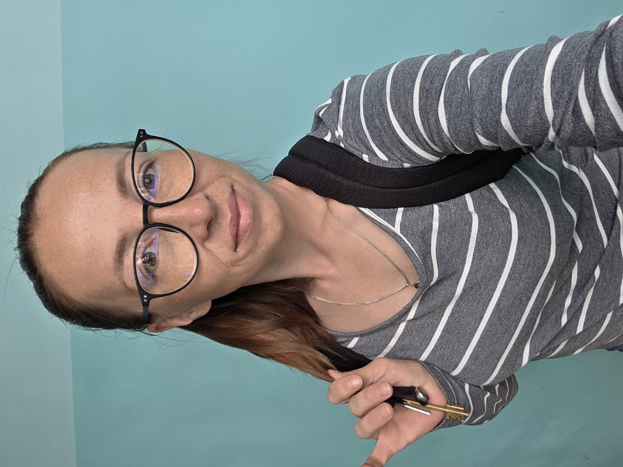 A woman with brown hair pulled back in a ponytail, wearing round black-rimmed glasses and a grey and white striped long-sleeved shirt, stands in front of a teal-colored wall. She is holding a set of keys in her left hand, which is resting on her shoulder, and has a black backpack on. She is looking towards the camera with a slight smile. The image appears to be a selfie taken indoors. There is no indication of the city of Chaikovsky, Russia, in the image itself or in the provided crops.