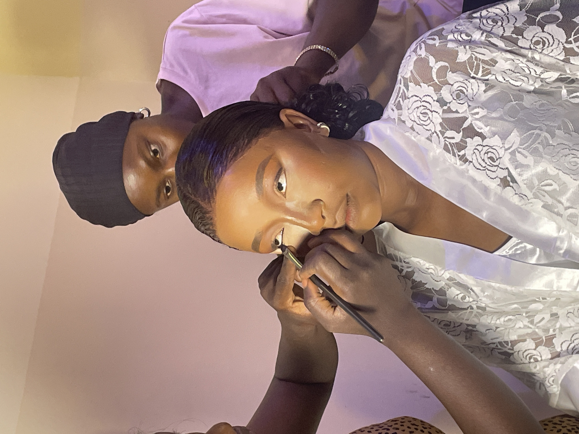 A medium shot captures a dark-skinned woman getting her makeup done. She is wearing a white lace robe and has short, styled hair. A makeup artist, wearing a black durag and a light pink shirt, is applying eyeliner to her right eye with a brush. The background is a soft, light pink. The location is unknown and Jos, Nigeria cannot be confirmed.