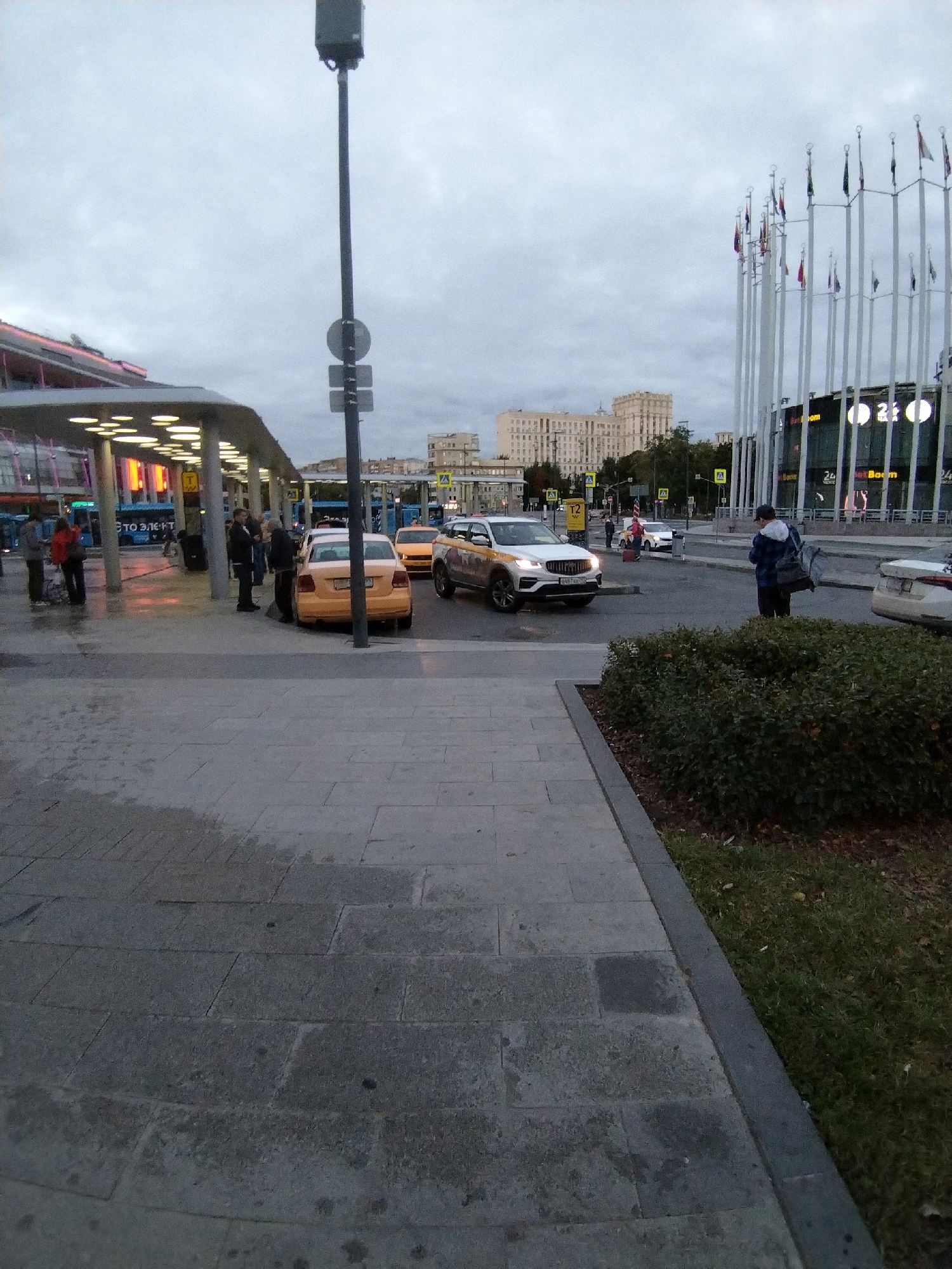 A street scene shows a pedestrian walkway with a covered waiting area, several yellow taxis, and a large building with many flags on poles in the background. The sky is overcast. In the foreground, the ground is paved with large rectangular stones, some of which are wet. To the left of the walkway, a covered structure with bright yellow lights illuminates a seating area. Several people are standing in and around this area, some appearing to be waiting.
Further down the walkway, a yellow sedan taxi is parked, and a white SUV taxi is in the street. A person is standing on the sidewalk near the white SUV, facing away from the camera. In the middle ground, a modern building with a glass facade is visible, adorned with numerous tall flagpoles. Two yellow taxi cars are present in the street. In the background, several multi-story buildings can be seen. The scene appears to be a transportation hub or a busy urban area.