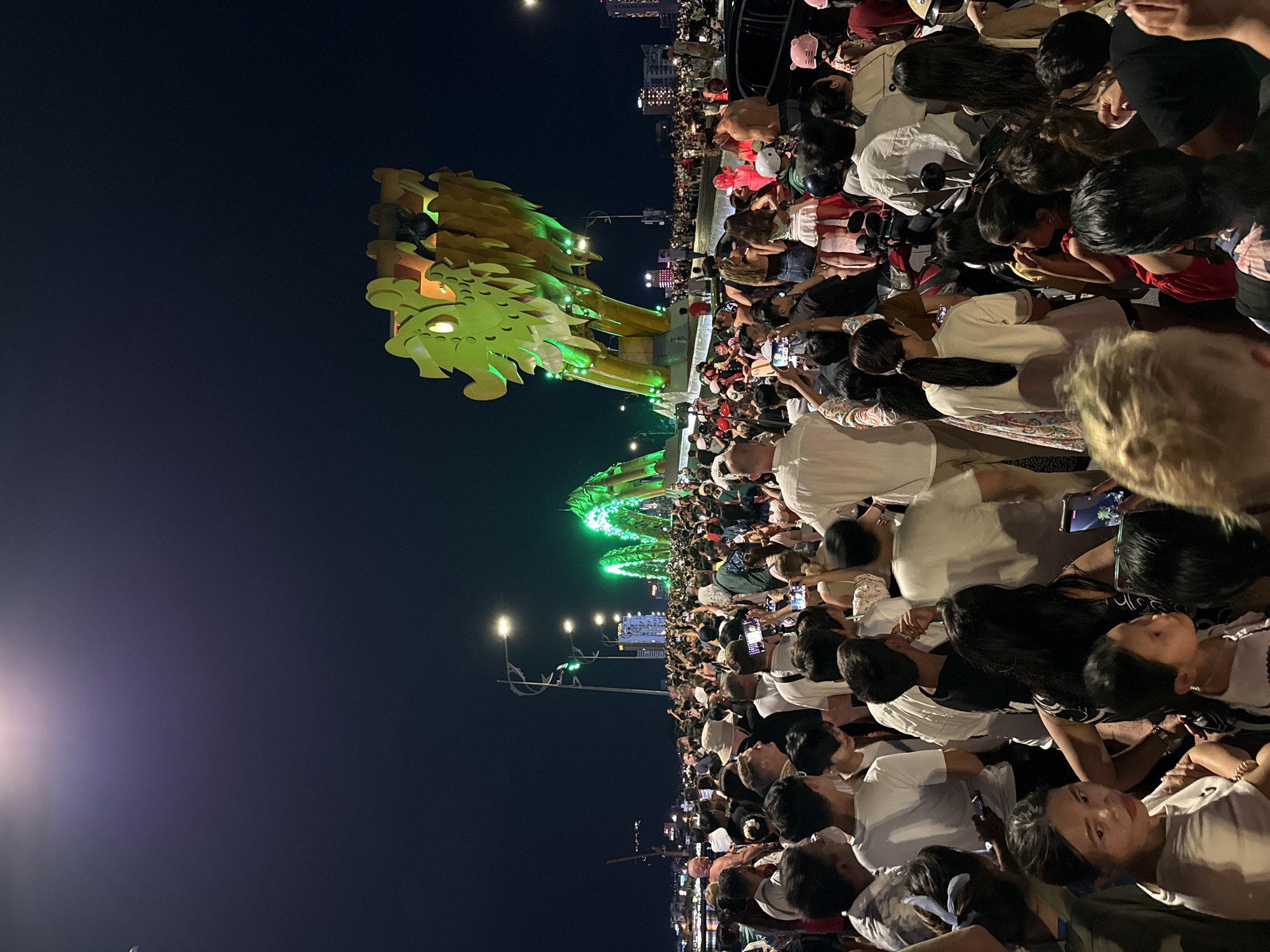 The image displays a dense crowd gathered on the Dragon Bridge (Cầu Rồng) in Đà Nẵng, Vietnam, at night. The bridge's main arches are illuminated with bright green lights, and the prominent yellow-green dragon head is visible on the right side. People in the foreground and midground are tightly packed, with numerous individuals holding smartphones elevated, directed towards the bridge structure. The crowd consists of adults and children, predominantly dressed in casual attire. Streetlights are visible along the bridge, providing additional illumination. In the background, urban buildings with illuminated windows and a distant construction crane are discernible across the river. Several motorbikes and scooters are interspersed among the people on the bridge. One man in the midground on the right is shirtless, standing near a parked black vehicle.