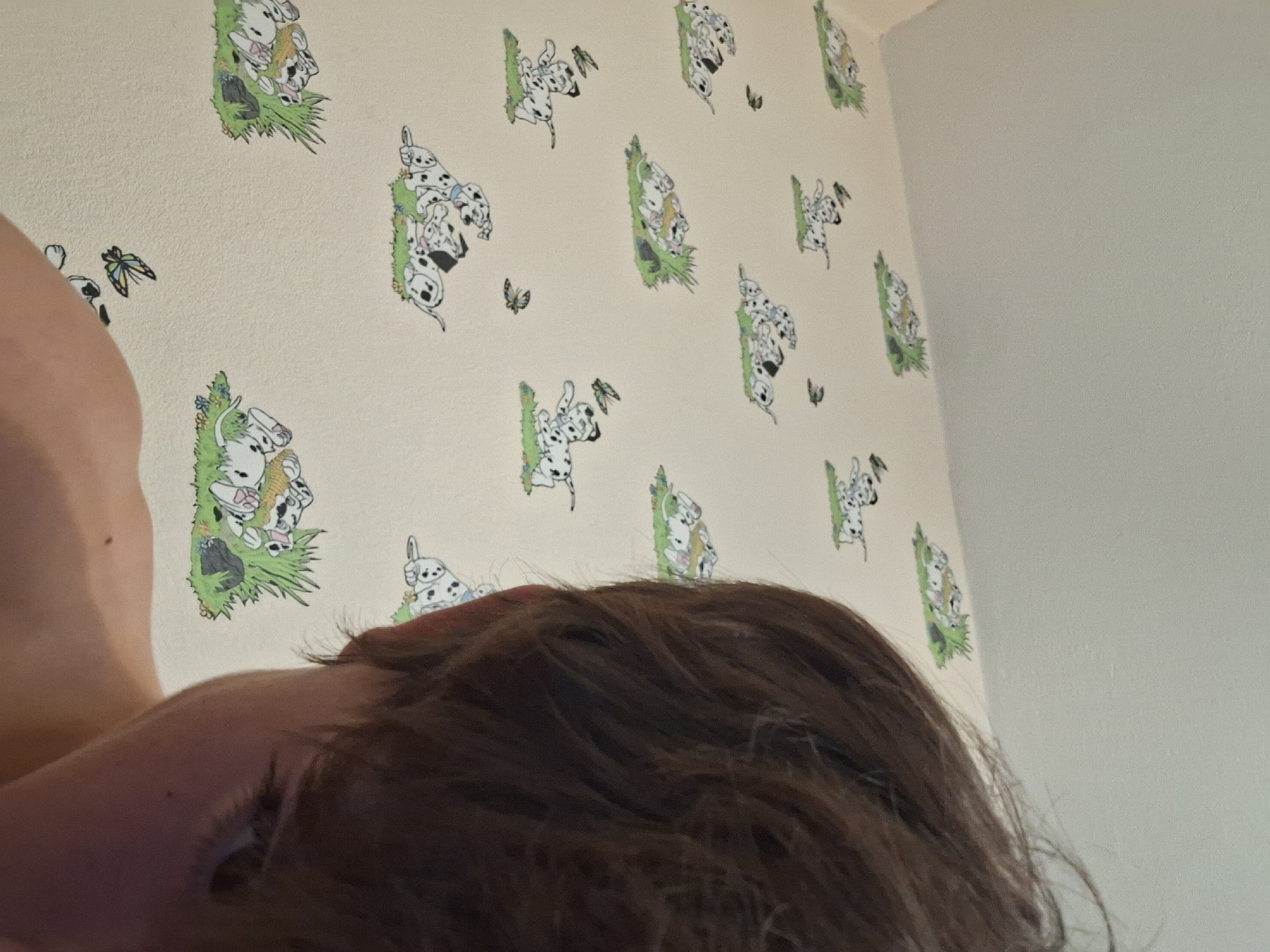 The image shows a close-up of a person's head and shoulder, partially obscuring a wall decorated with wallpaper featuring a repeating pattern of cartoon Dalmatian puppies. The wallpaper has a light cream background and depicts various scenes of puppies playing and resting on green grass, with some small black dots or birds scattered around. The person has brown hair and is looking down and to the right, with their right eye visible. The lighting suggests an indoor environment. The image was taken in Varna, Bulgaria.