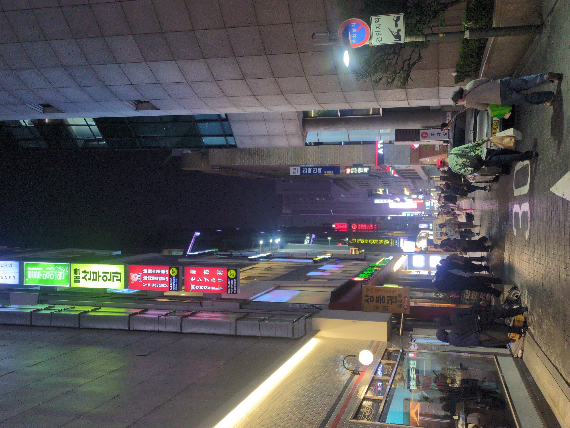 This is a street scene in Seoul, South Korea at night. A building with a parking restriction sign is on the right side of the frame, and several people are unloading items from a silver vehicle. Further down the street, a crowd of people are walking on the sidewalk. Neon signs in Korean and English adorn the buildings, advertising businesses such as restaurants ("Beef Buffet") and medical clinics. There's a sign for "Mongvely" (transliterated from Korean). Streetlights illuminate the scene, and reflections are visible in the shop windows.