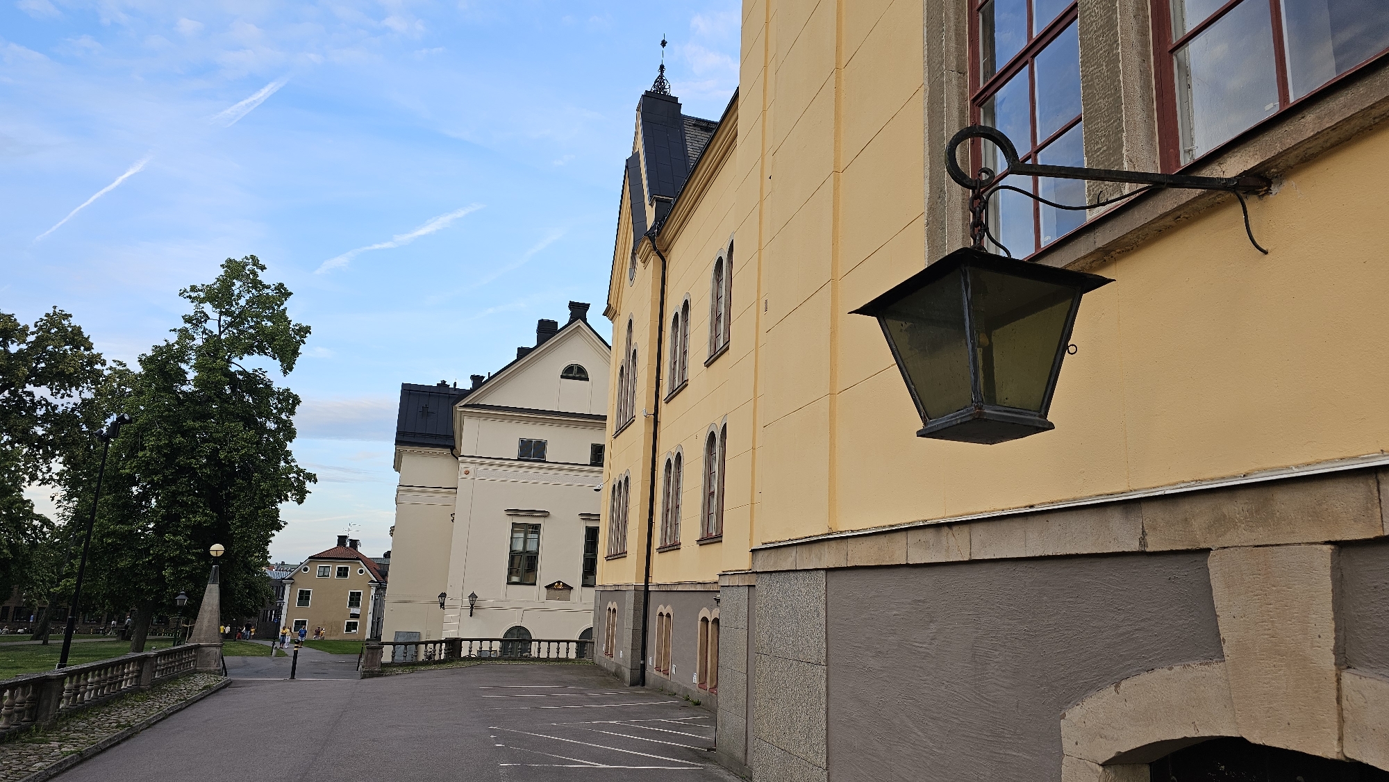 This image captures a side view of a yellow building with a grey stone base, featuring a dark, antique-style lantern hanging from an ornate metal bracket on the exterior wall. The building has several arched windows with dark frames, and the wall appears to be stuccoed. Behind this building, another white building with a dark roof is visible, also with arched windows. Further in the background, a lighter colored building with a red roof and multiple windows is visible, along with a park area featuring large, mature trees and people milling about. The sky is blue with wispy white clouds, and the presence of contrails suggests an airplane has recently passed by. The lighting indicates it is likely late afternoon or early evening. The scene appears to be a public space, possibly a historical district or a university campus, given the architectural style and the presence of a park. No text is visible in the image.