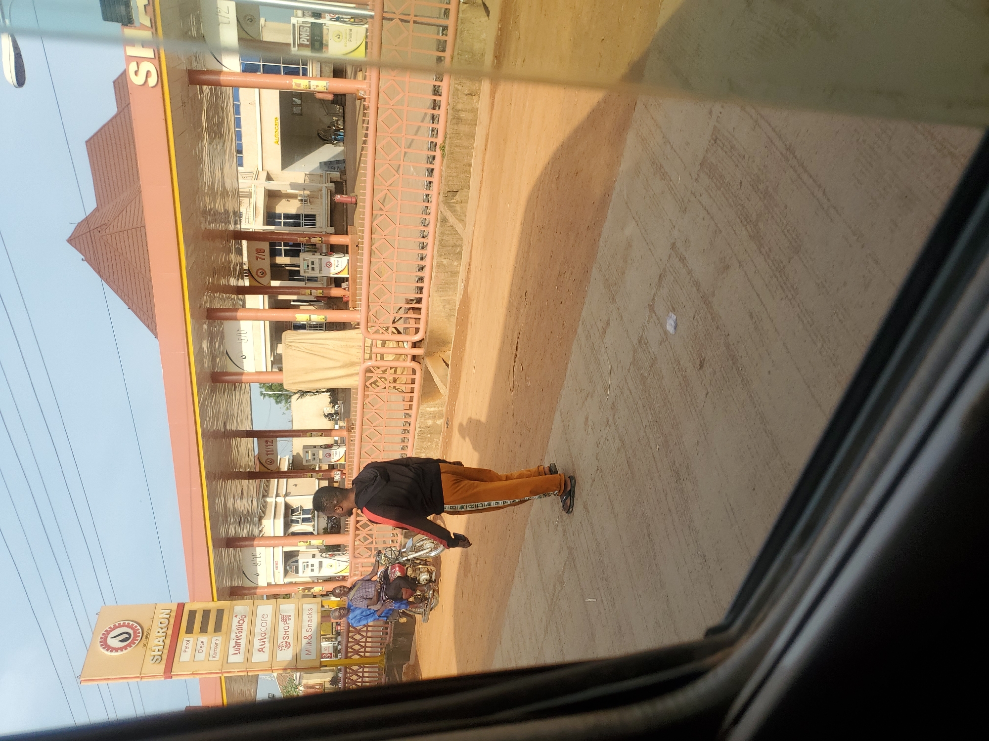 The image shows a street scene in Kakau, Nigeria, featuring a SHARON gas station. The gas station has a prominent sign with the brand name and categories like Petrol, Diesel, Kerosene, Lubricants, Autocare, Shop, and Milk & Snacks. Several fuel pumps are visible under the covered area of the station.
In the foreground, a man wearing a black hoodie and orange pants with black stripes is standing facing away from the camera. His posture suggests he is either waiting or observing something. To his left, on a motorcycle, are two men. The man in the front is wearing a blue shirt, and the man behind him is wearing a checkered shirt. They appear to be interacting or waiting.
The ground is a mix of paved concrete and dirt. The overall environment suggests an outdoor, possibly commercial area. The lighting indicates it's daytime, with shadows cast by the structures and people. The view appears to be from inside a vehicle, as evidenced by the blurred foreground elements suggesting a window.