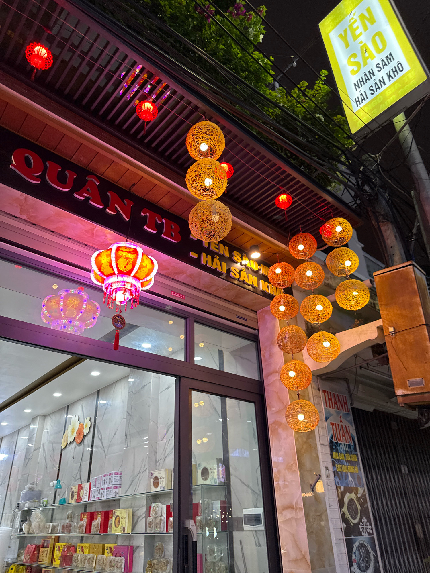 This image captures a street scene at night in Nha Trang, Vietnam. The foreground features a store with a large glass window displaying shelves stocked with various packaged goods, including boxes that appear to contain food items or gifts. Above the store, a prominent red sign with white lettering reads "QUÂN TB." Suspended from above are strings of decorative lanterns, primarily spherical and made of woven material in shades of yellow and orange, casting a warm glow. Smaller red lanterns are also visible. To the upper right, a bright yellow sign with Vietnamese text, "YẾN SAO NHÂN SÂM HẢI SẢN KHÔ," is illuminated. The environment suggests a commercial street, with electrical wires overhead and a glimpse of foliage from trees. The overall impression is a vibrant and festive nighttime atmosphere, typical of a commercial area in Vietnam.