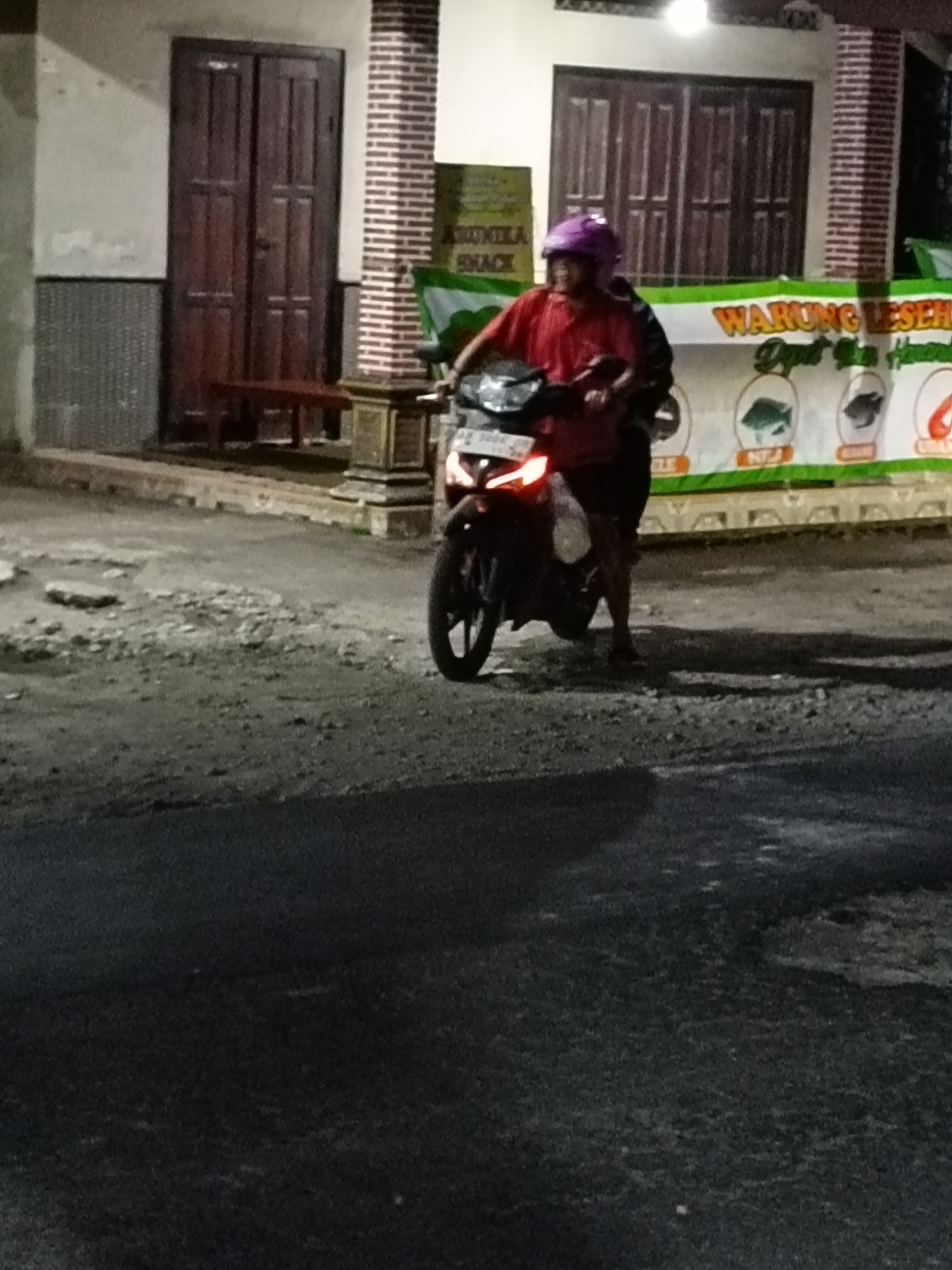 A person wearing a purple helmet and a red shirt is riding a motorcycle on a dirt road. The motorcycle is black and red and has its headlights on. In the background, there is a building with a large wooden door and a sign that says "WARUNG LESEH". There are also some potted plants and a small table in front of the building. The scene appears to be in a residential area.