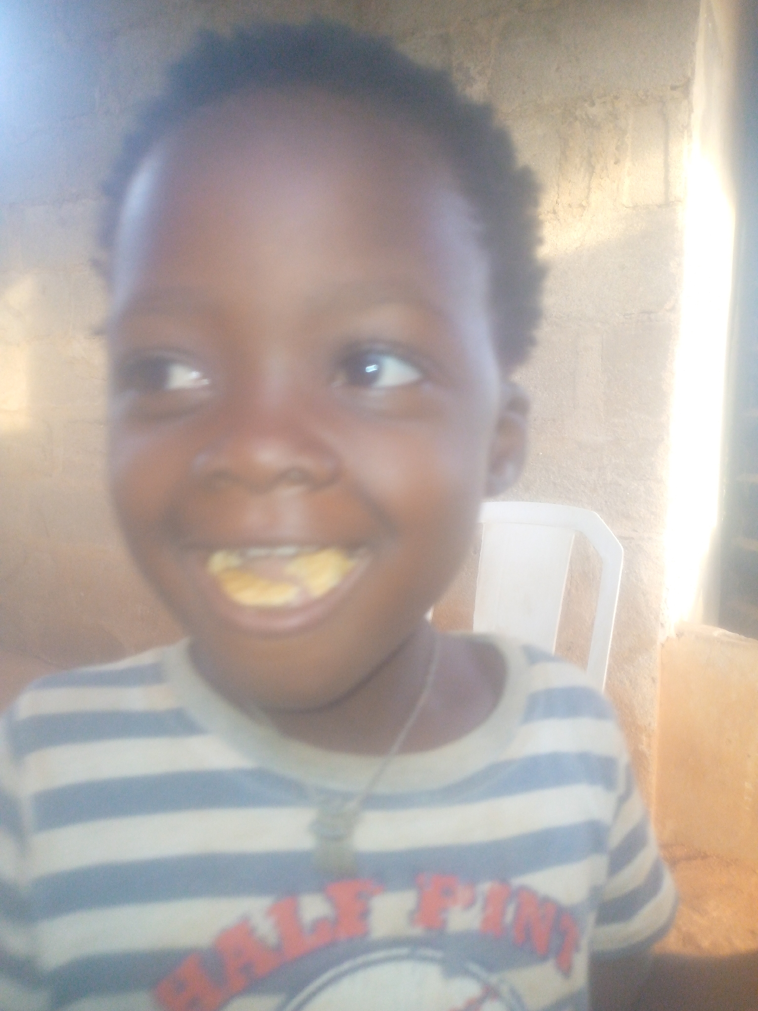 The image shows a young child with dark skin and short, dark hair. The child is wearing a striped shirt with a possible inscription on the front, though the details are unclear. The child has something in their mouth, appearing to be food.
In the background, there is a white plastic chair and a wall that seems to be made of stone or brick. Bright light is coming from the right side of the frame. The location is Unknown, Unknown.