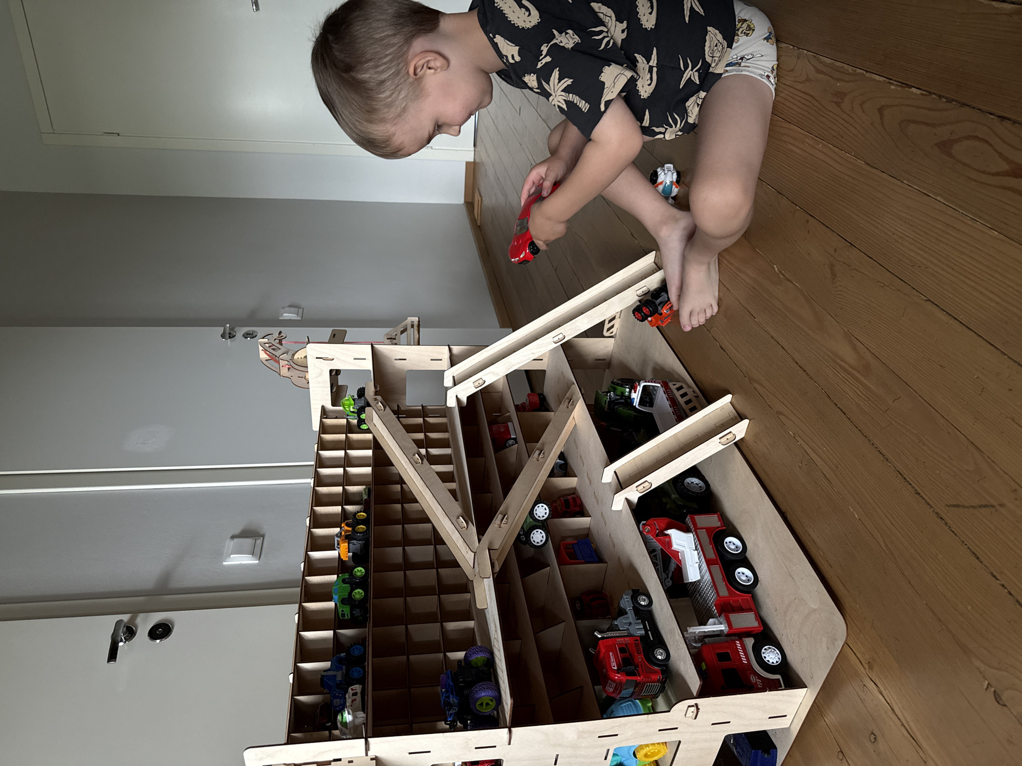 In the city of Chaikovsky, Russia, a young boy with light brown hair is playing with a wooden toy garage and toy cars on a wooden floor. The boy is sitting on his right side, looking towards the toy garage. He is wearing a black t-shirt with a white pattern and white shorts with cartoon characters. In his right hand, he holds a red toy car. Several toy cars of various types, including monster trucks, a fire truck, and a tractor, are placed in the compartments and on the ramps of the multi-level wooden toy garage. The garage is made of light-colored plywood and has ramps that connect different levels. In the background, a light-colored door with a silver handle is visible. The lighting suggests an indoor environment.