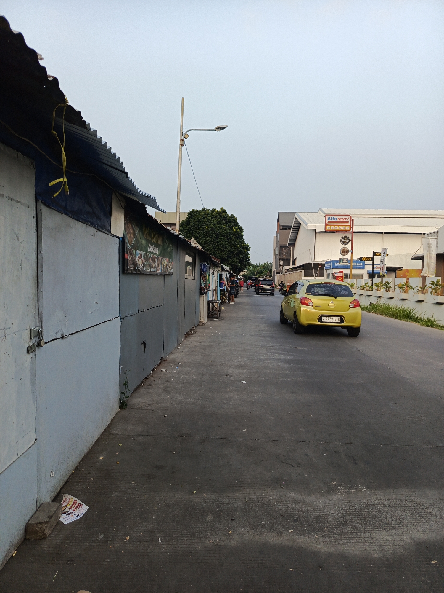 This is a vertical, eye-level shot of a street in Tangerang, Indonesia, with a focus on the roadside structures and a yellow car on the road. On the left side of the image, a row of small, dilapidated roadside stalls with corrugated metal roofs and light blue-grey walls stretches into the distance. A banner with images of food is displayed on one of the stalls. The street itself is paved with asphalt and shows signs of wear, with debris scattered along the edge and on the sidewalk. A yellow car is in the right lane of the road, heading away from the viewer. In the background, a modern building with an "Alfamart" sign is visible. The sky is overcast and light blue.