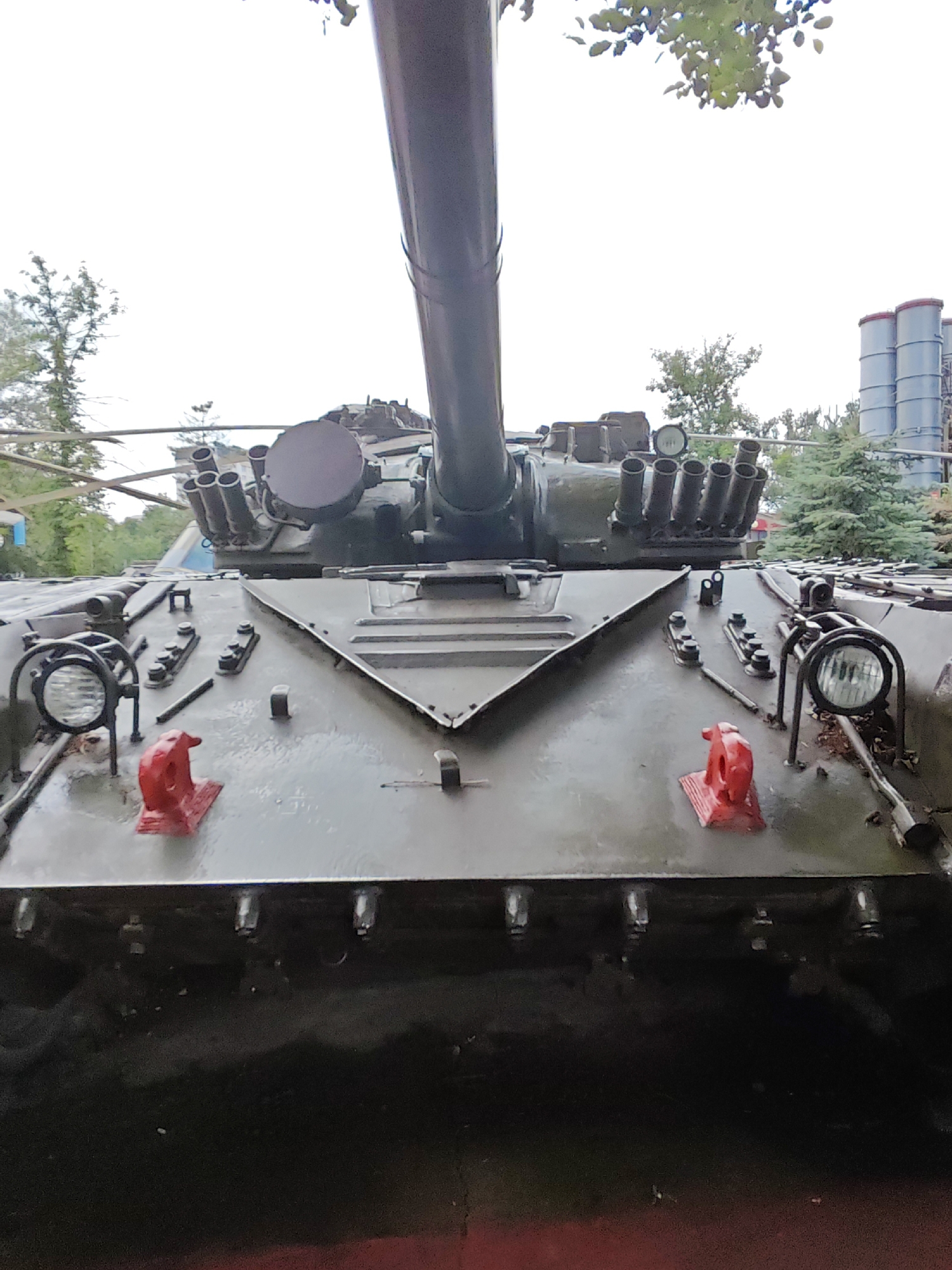 The image displays the front of a military tank. The tank is painted a dark green, and its main cannon extends upwards and out of frame. The front of the tank features various details, including two circular headlights with protective grilles, two bright red tow hooks, and what appear to be smoke grenade launchers or other weapon systems mounted in front of the turret. The tank's surface appears wet, suggesting recent rain or washing. In the background, trees and foliage are visible, with what looks like industrial structures or silos in the far distance. The overall impression is that of a static display, possibly in a museum or park, given the context of its likely location in Cherkessk, Russia, where such military hardware is often displayed. There are no people visible in the image.