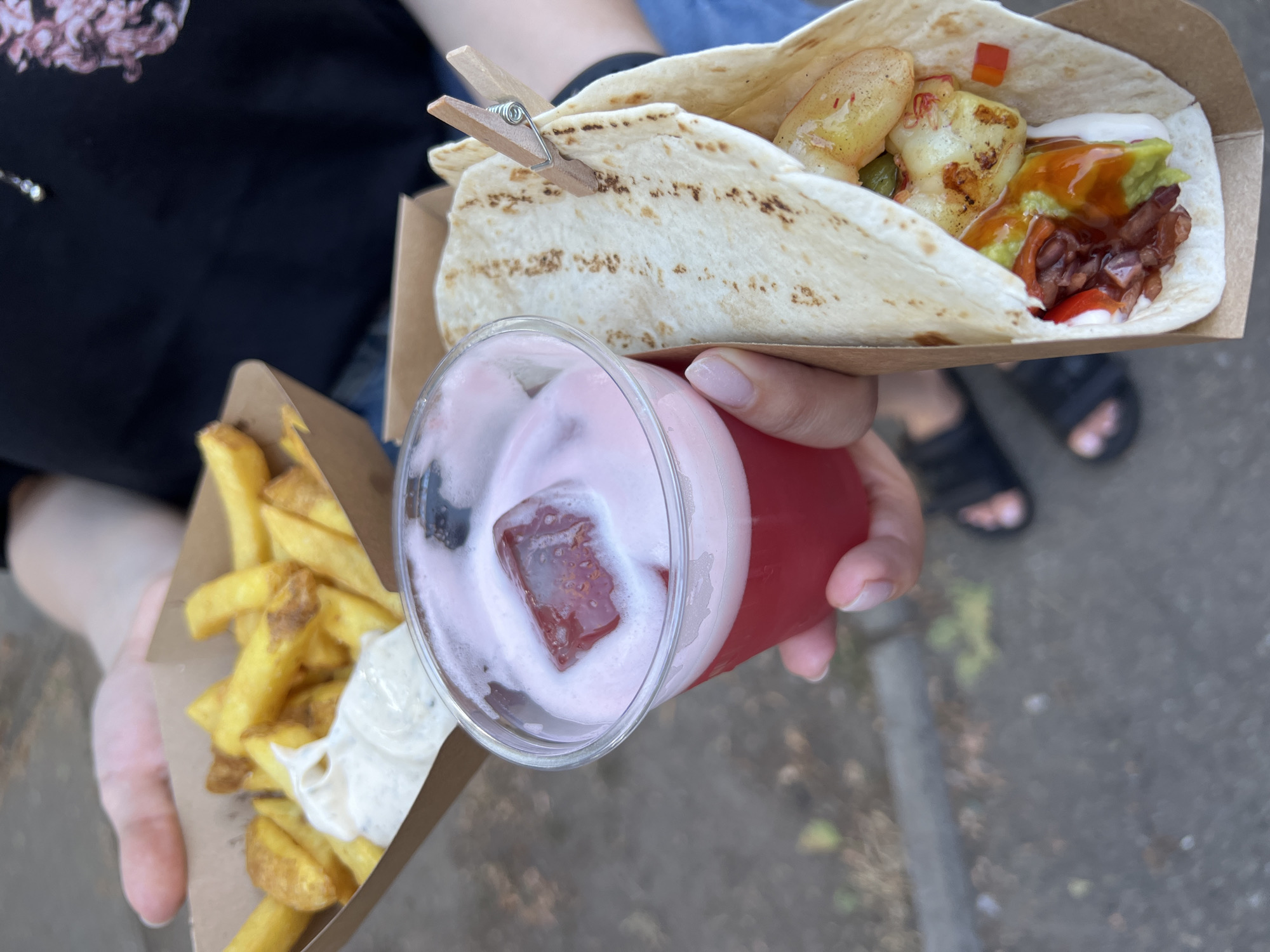 In Kyiv, Ukraine, two hands hold takeaway food items: a carton of french fries with a dollop of white sauce and a clear plastic cup of a frothy pink beverage with a single red ice cube. Beside these, a flatbread is folded around various fillings, secured with a wooden clothespin. The background suggests an outdoor urban setting, possibly a street or park, with visible pavement and some greenery. The food appears to be consumed on the go.
