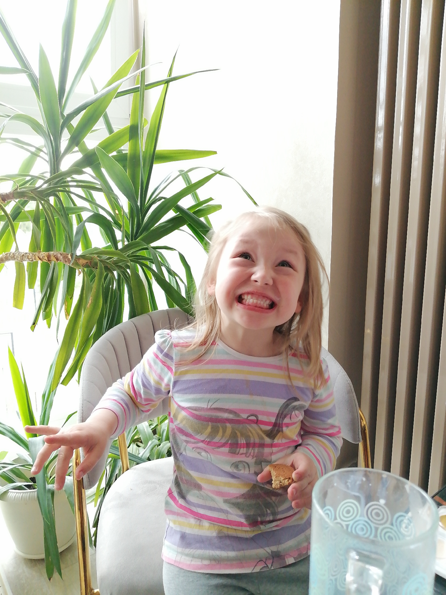 A young, fair-skinned child with blonde hair sits in a grey chair with gold legs. The child wears a long-sleeved, striped shirt and holds a piece of food in one hand. The child is smiling widely, displaying their teeth. Behind the child is a large potted plant with long, green leaves. To the right of the child is a grey radiator. In the foreground, there is a glass with a patterned design. The setting appears to be indoors, possibly in a home or apartment. There is no information in the image to confirm a location in the city of Trubacheyevka, Russia.
