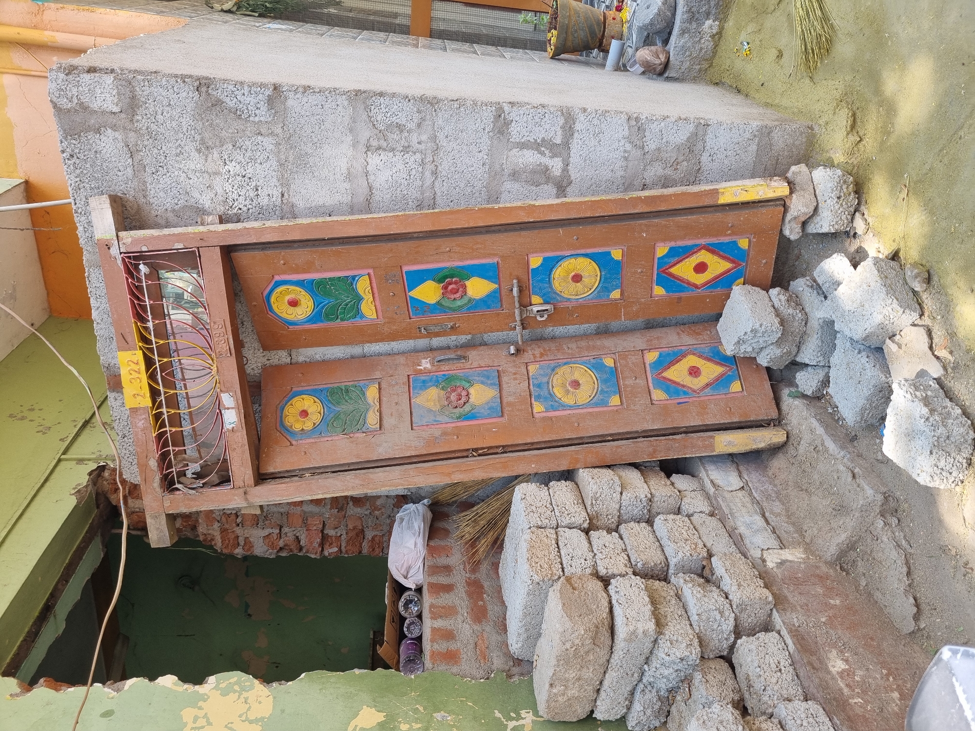 An outdoor scene in Pedda Kammavaripalli, India, depicts a construction or renovation site. A detached brown wooden double door frame, featuring an arched wrought-iron grille with yellow, red, and orange concentric rings, is propped upright. The door's panels are adorned with colorful, faded, painted designs in yellow, red, blue, and green, depicting flowers and geometric shapes. A silver-colored metal latch mechanism is visible on the right door panel. The door rests on several rough gray concrete blocks on the ground, leaning against two distinct wall structures.
To the left, a section of wall painted faded green-yellow exhibits significant peeling, revealing reddish-brown brickwork. An open doorway or large aperture in this wall exposes an interior cavity with exposed brick. On a narrow ledge within this opening, three rolls of material (one purple, two white with patterns) and a white plastic bag are visible. Below this, a stack of approximately 14-16 rectangular gray concrete blocks is neatly arranged. A natural fiber broom leans against the wall nearby.
To the right of the door, a wall constructed from gray concrete blocks, some textured and some with a smoother light gray finish, is visible. At the top of the door frame, a yellow sign with the inscription "2-322" is affixed. The ground in the foreground consists of dirt, with patches of fine yellow powder, possibly construction dust or cultural powder. Scattered small concrete fragments lie near the base of the door. Further to the right, a dark brown pot with yellow contents, possibly flowers or petals, sits on an elevated surface. No individuals are present in the scene. The area is brightly lit by daylight.