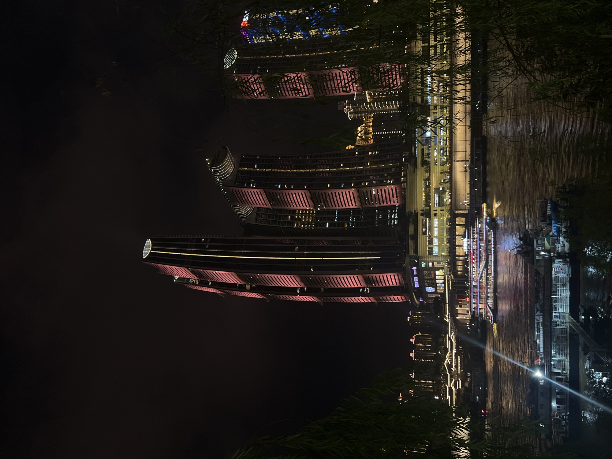 A night view of the Jiangbeicheng Street in Chongqing, China, shows illuminated skyscrapers and a river with boats. The scene is dominated by the striking architecture of the Raffles City Chongqing complex, with its distinctive curved towers illuminated in pink and yellow lights. Other buildings in the background are also lit up, creating a vibrant urban landscape against the dark night sky. The Yangtze River flows in the foreground, with several boats of various sizes docked or moving on its surface. One large passenger boat, adorned with bright lights, is prominently featured. The foreground is partially obscured by the dark foliage of trees, adding depth and a touch of nature to the urban setting. The overall atmosphere is one of modern urban development and nighttime activity.