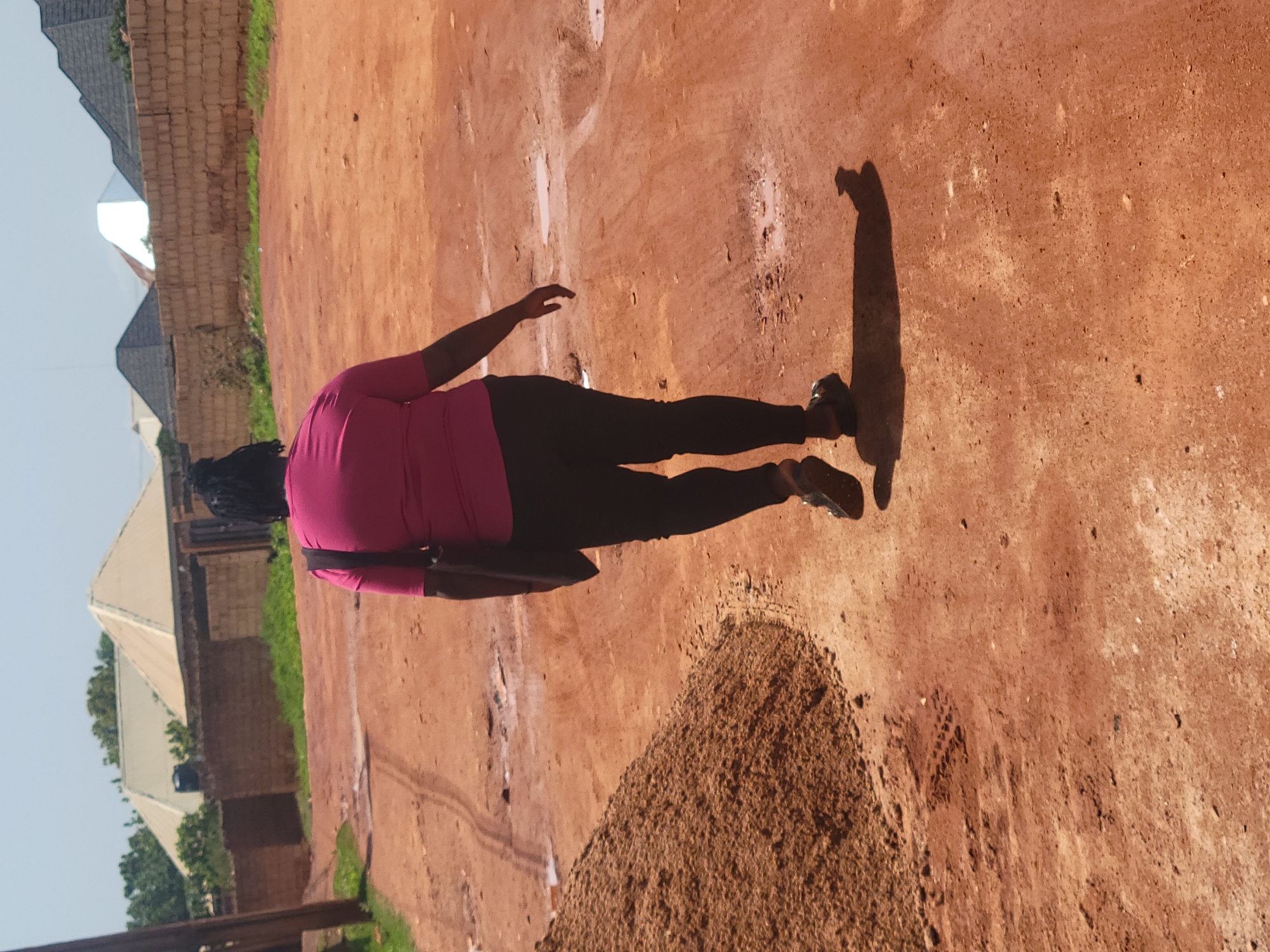 The image depicts a woman walking away from the camera on a dirt road in the city of Jos, Nigeria. She is wearing a bright pink t-shirt and black leggings, with a dark bag slung over her shoulder. Her hair is styled in braids. The road is uneven with puddles of water and tire tracks, indicating it may be a less developed area. To the left of the road, a large pile of sand is visible. In the background, several buildings with distinct architectural styles, including what appear to be newly constructed houses with corrugated iron roofs, are seen behind a low brick wall. The sky is clear and blue, suggesting a sunny day.