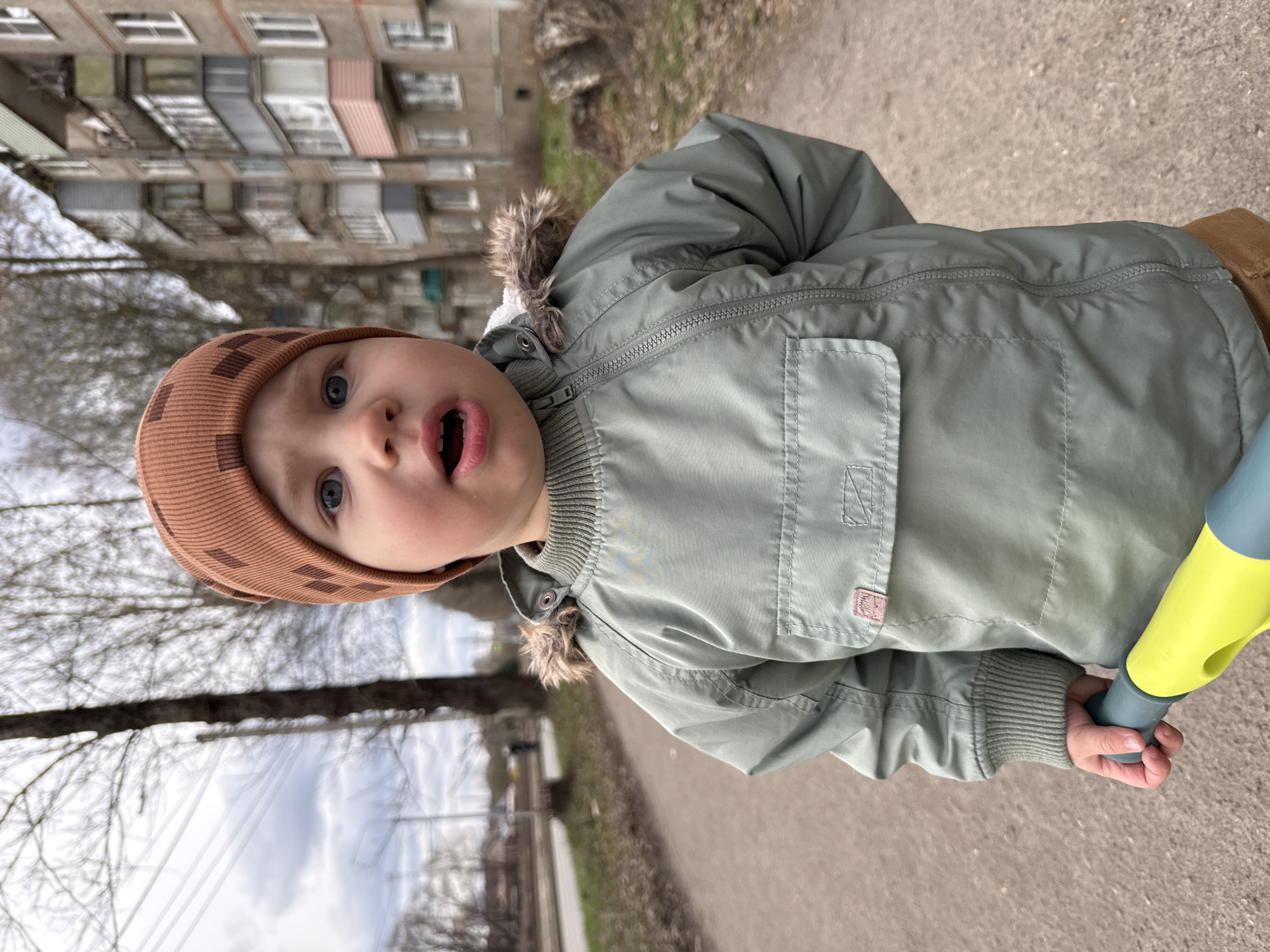 A young child stands on a paved path, likely sidewalk, in an urban environment. The child wears a brown knit hat and a green jacket with a fur-trimmed hood. The mouth is open. The child holds a yellow and blue object, possibly a toy. A multi-story building is visible in the background. Bare trees line the path. Based on the architecture of the building and the overcast sky, the location is likely Zhukovo, Russia.