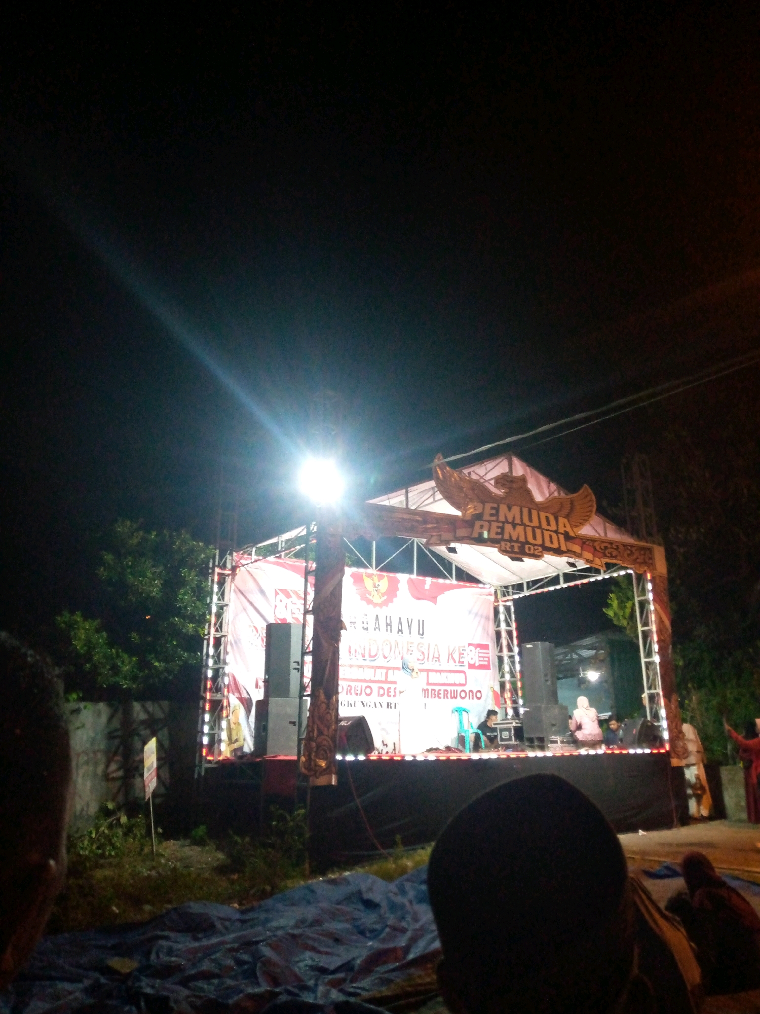 This is a nighttime, exterior, ground-level, medium-distance shot of an outdoor stage event in Sumberwono, Indonesia. The stage is illuminated by bright lights, with a large banner that reads "PEMUDA REMUDI RT 02" and "DIRGAHAYU INDONESIA KE 78". A large golden eagle emblem is affixed to the top of the stage structure. Several speakers are visible on and around the stage, along with a blue chair and what appears to be a person dressed in a yellow outfit sitting on the stage. The foreground shows a dark tarp spread on the ground and the backs of several people's heads, suggesting an audience. Trees and a wall are visible behind the stage.