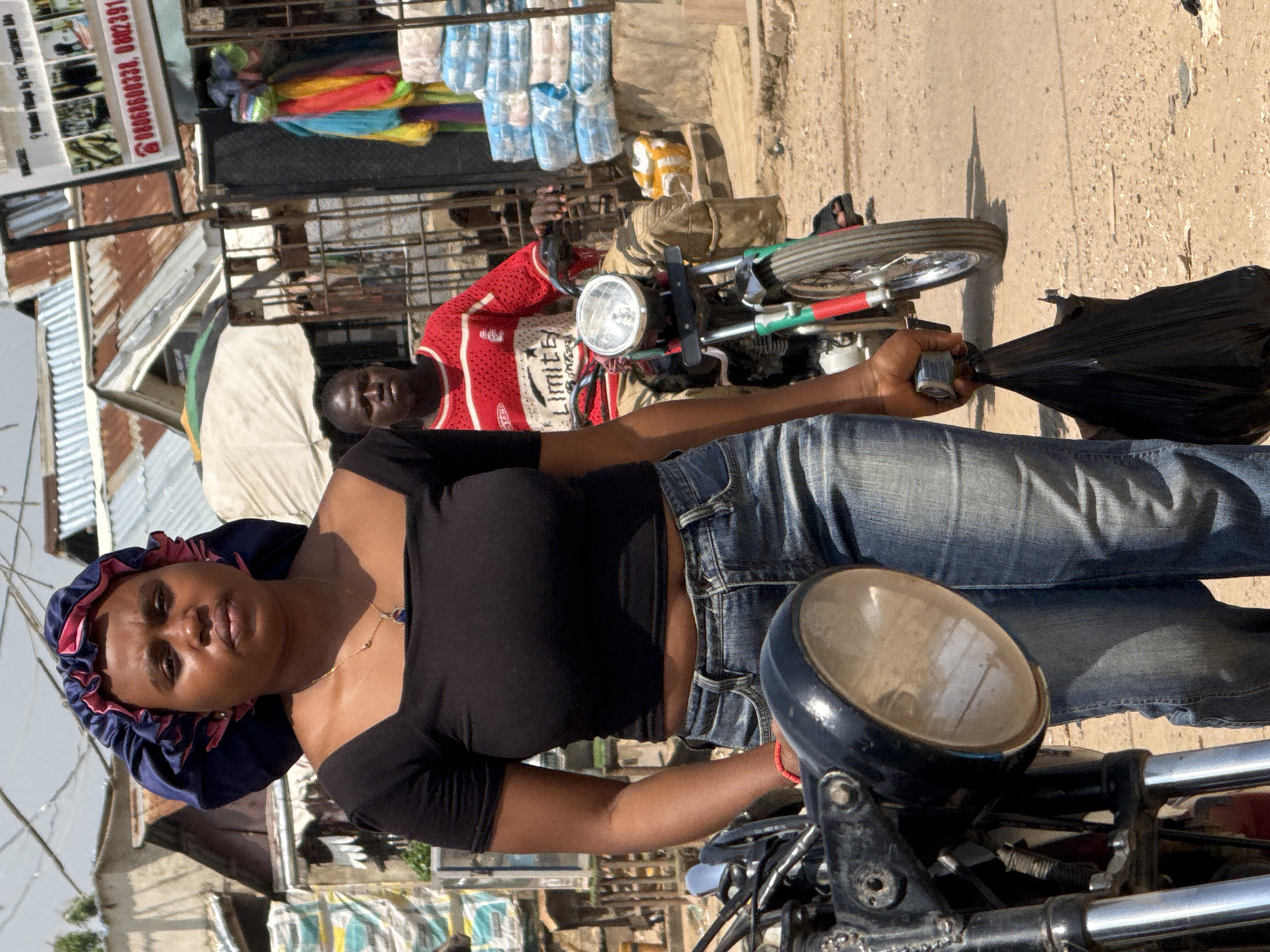 A woman with a blue and pink bonnet walks in front of a motorcycle and rider on a dirt road in Abuja, Nigeria. She wears a black crop top and blue jeans, holding a black plastic bag in her right hand. The man on the motorcycle wears a red and white striped long-sleeved shirt and brown pants. The background shows buildings with corrugated metal roofs, stalls with colorful textiles, and stacks of packaged goods. The scene appears to be a busy marketplace or roadside area.