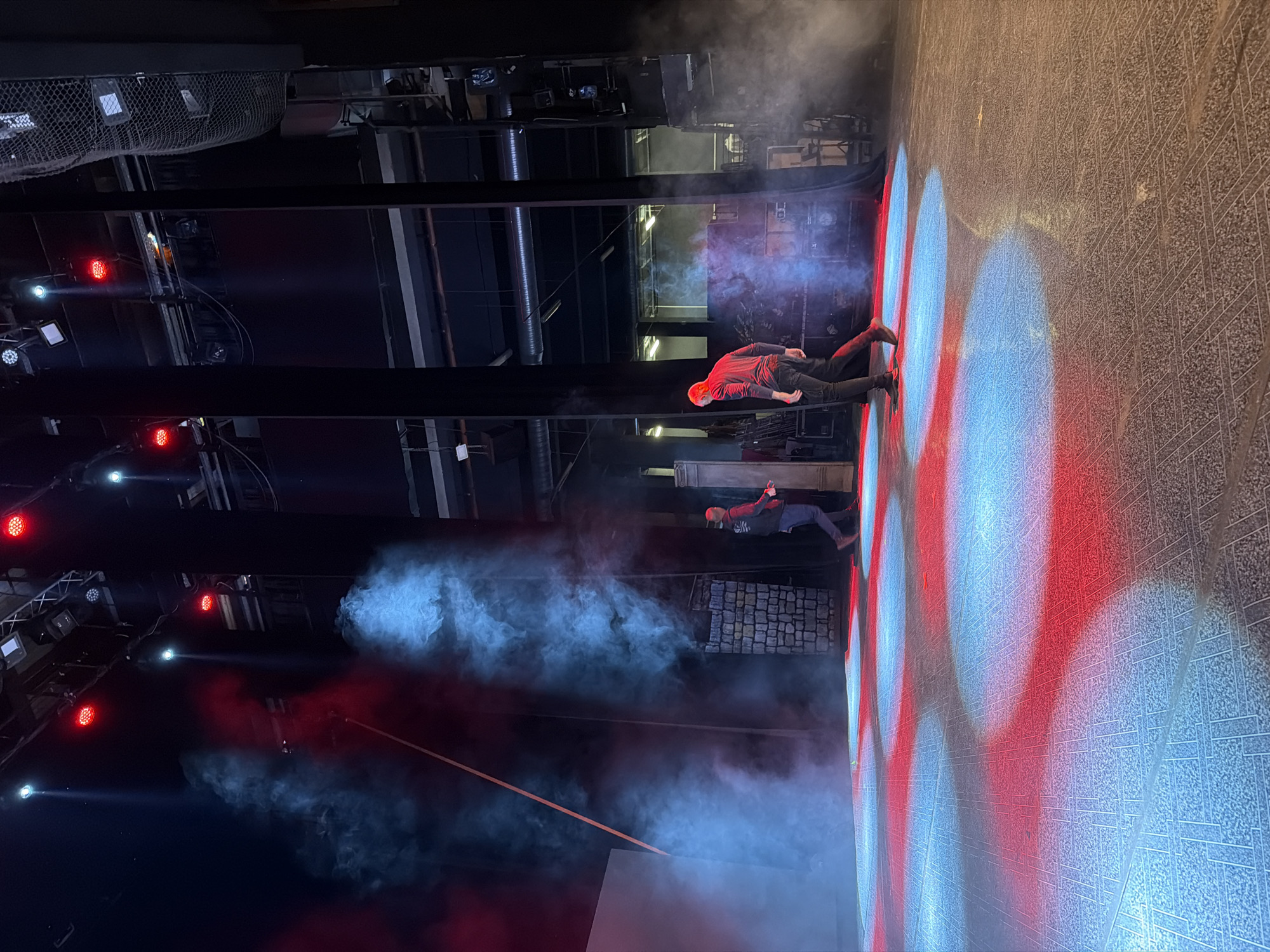 The image depicts a theatrical performance in Plovdiv, Bulgaria. Two men wearing dark pants and red shirts are positioned on a brightly lit stage with a red and blue pattern. Smoke fills the space around them. The stage is set within a larger structure with visible lighting rigs and support beams.