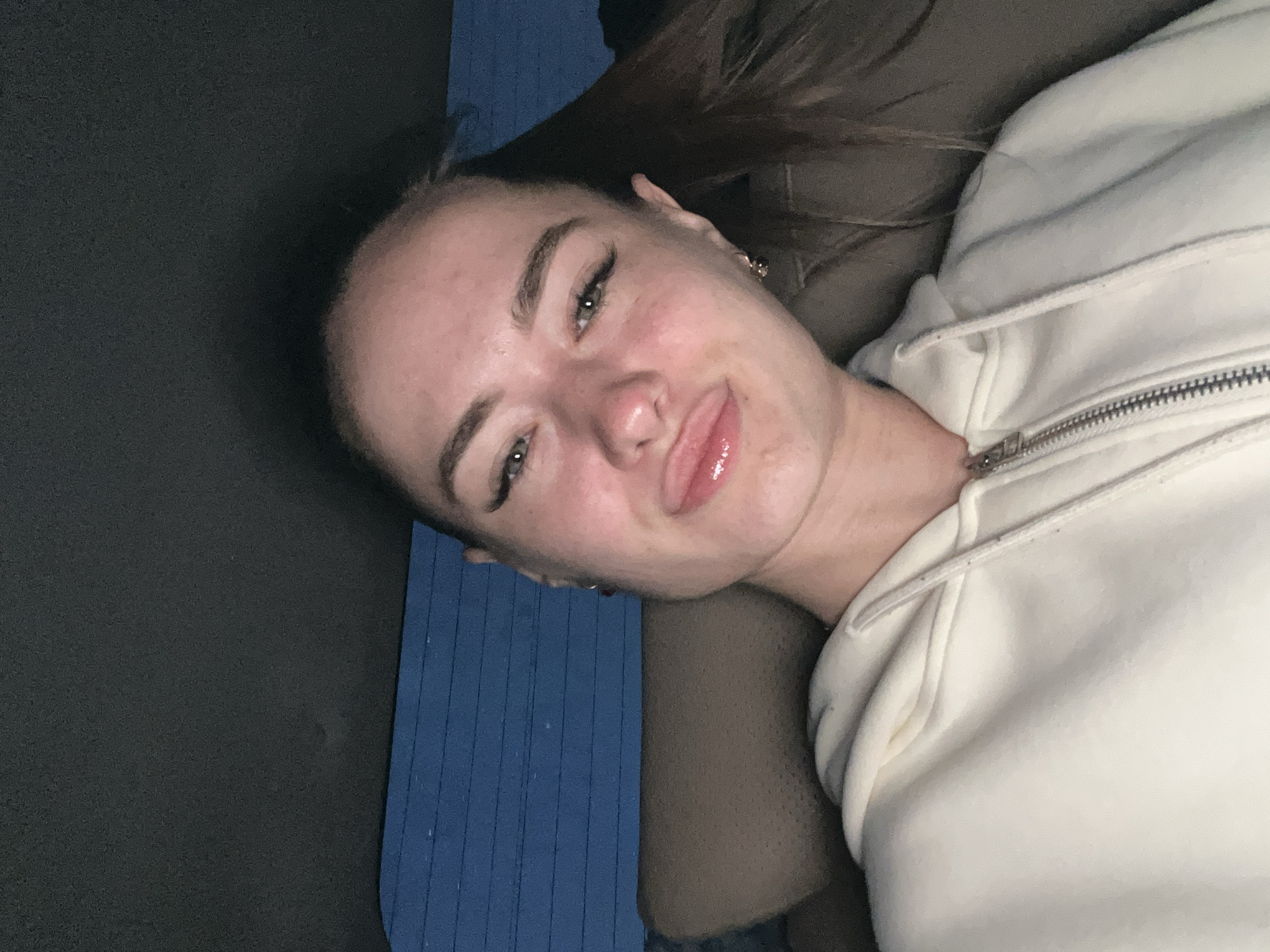 The media file is a close-up, self-taken photograph of a young woman. She is lying down, with her head tilted, and her face takes up a significant portion of the frame. She has fair skin, light-colored eyes, and dark eyebrows, with noticeable eyelashes. Her lips are slightly parted, and she is looking directly at the camera. She is wearing a light-colored hooded sweatshirt, partially zipped. Her hair is dark and falls behind her.
The background is dark and out of focus, with some hints of blue, possibly a mat or furniture, and a darker, textured surface to her left. The lighting suggests an indoor setting, possibly at night or in a dimly lit room, as there are no strong light sources visible and the shadows are soft. Her expression is relaxed and casual, possibly conveying a sense of contentment or playfulness. There is no visible text in the image. The overall impression is that of a candid, personal photograph.
