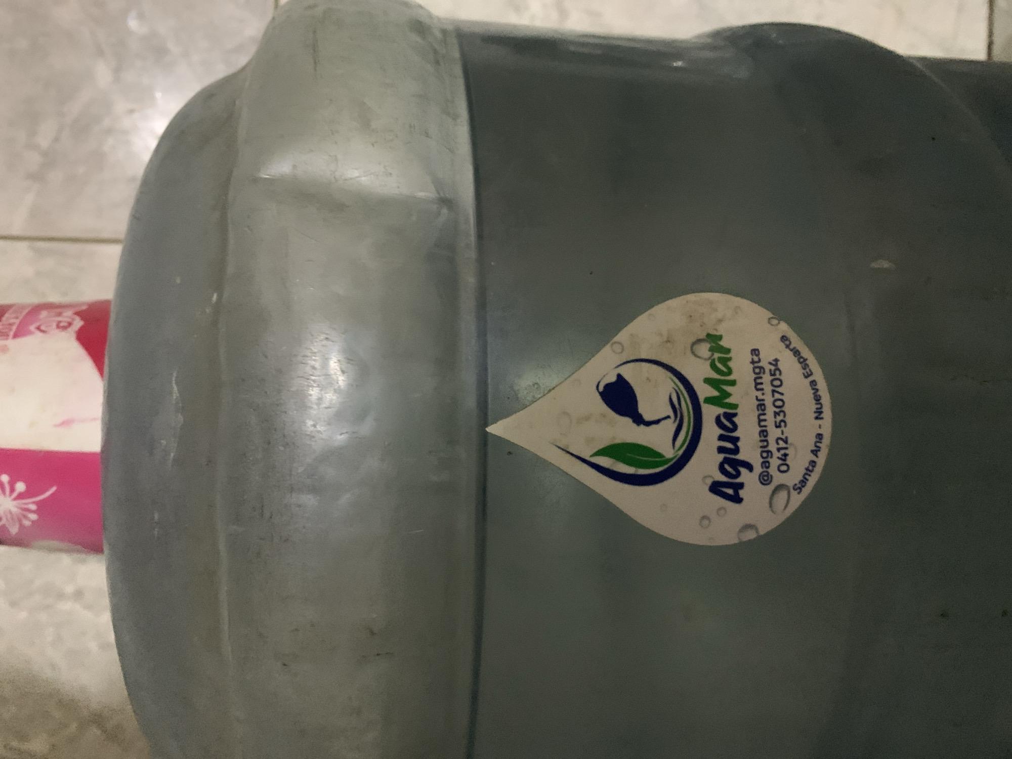The image presents a close-up view of a large, translucent blue plastic water bottle, typically used for water dispensers. The bottle is lying horizontally on a light-colored, possibly tiled floor. A portion of a pink object with a white floral design is visible on the left side of the frame.
A white, teardrop-shaped label is affixed to the side of the water bottle. The label features a logo with a blue, stylized figure appearing to pour water from a jug, surrounded by a green leaf, all within a blue circle. The brand name "AquaMar" is prominently displayed in blue, stylized text. Below the brand name, smaller text provides contact and location information: "@aguamar.mgta", a phone number "0412-5307054", and "Santa Ana - Nueva Esparta," which confirms the location in Santa Ana, Venezuela.
The bottle shows signs of use, with some minor scuffs on its surface. The setting appears to be indoors, under ambient lighting, and there are no people or specific activities taking place.