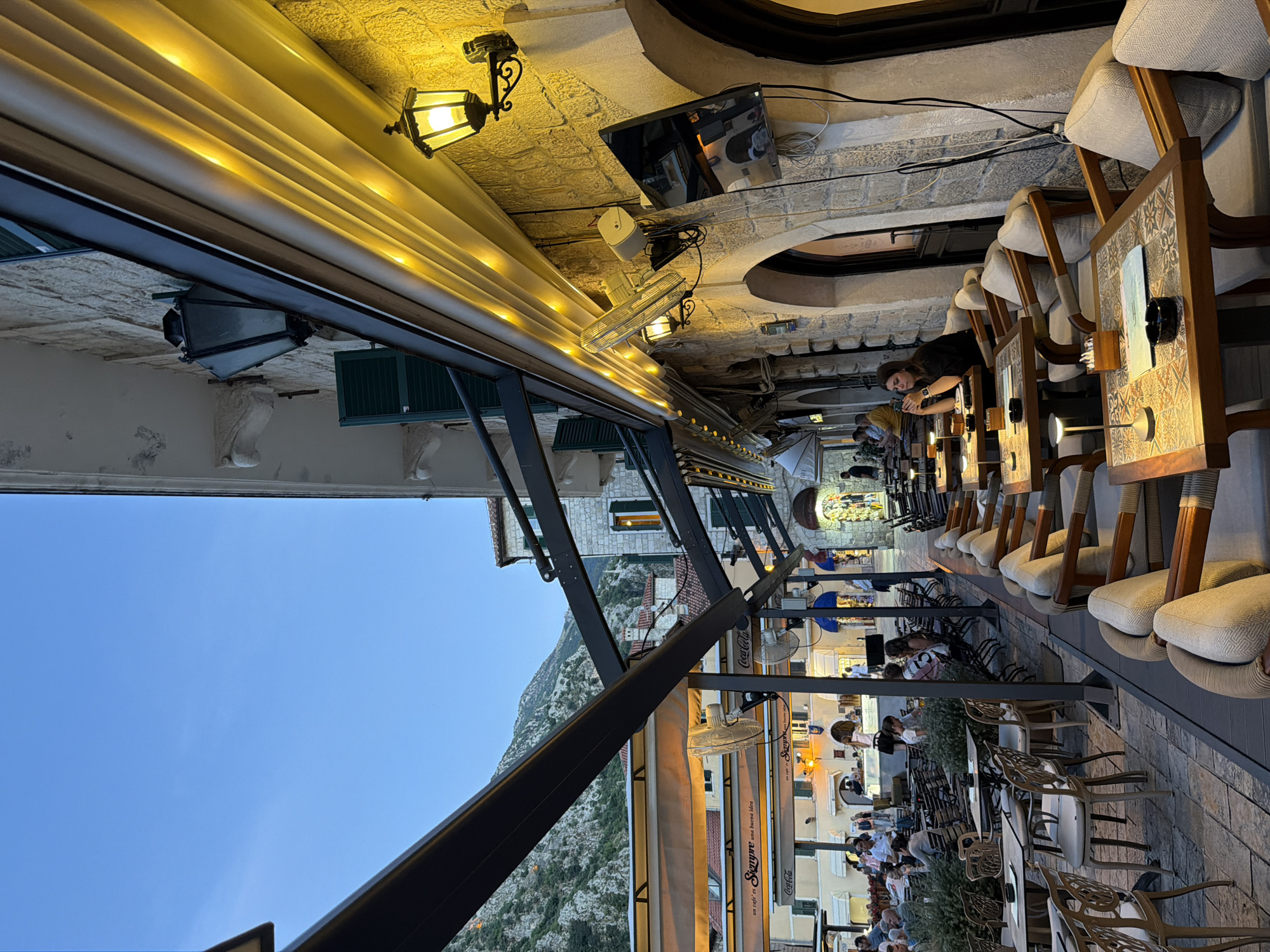 The image depicts an outdoor dining area in Kotor, Montenegro, likely at dusk or early evening. The scene is set along a cobblestone street lined with restaurants. In the foreground, several tables with chairs are arranged, some with table lamps and ashtrays. The architecture features stone buildings with arched doorways and windows. Warm yellow lights illuminate the facade of one building, casting a glow on the outdoor seating area. A woman in a black top is seated at one of the tables, looking towards the camera. In the background, more tables are visible, along with other patrons and the street stretching into the distance. Mountains are visible in the upper left portion of the image, suggesting the mountainous terrain surrounding Kotor. Signs for "Coca-Cola" and "Siempre" are visible on awnings, indicating the presence of cafes or restaurants. The overall atmosphere is that of a lively evening scene in a European city.