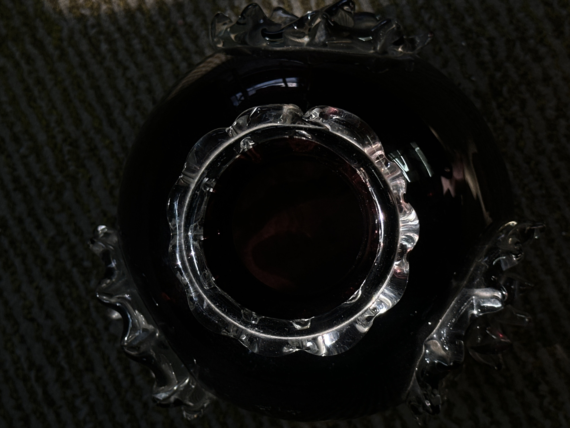 The image shows a close-up, top-down view of a dark, possibly purple, glass vase. The vase has a ruffled or scalloped rim made of clear glass. The interior of the vase is dark and reflective, with some reddish hues visible. The vase is resting on a textured surface that appears to be a rug or carpet. The lighting is dramatic, casting deep shadows and highlighting the glass elements. There are no people or specific landmarks visible. The image does not contain any information about the city of Luhansk, Ukraine, or any other geographical location.