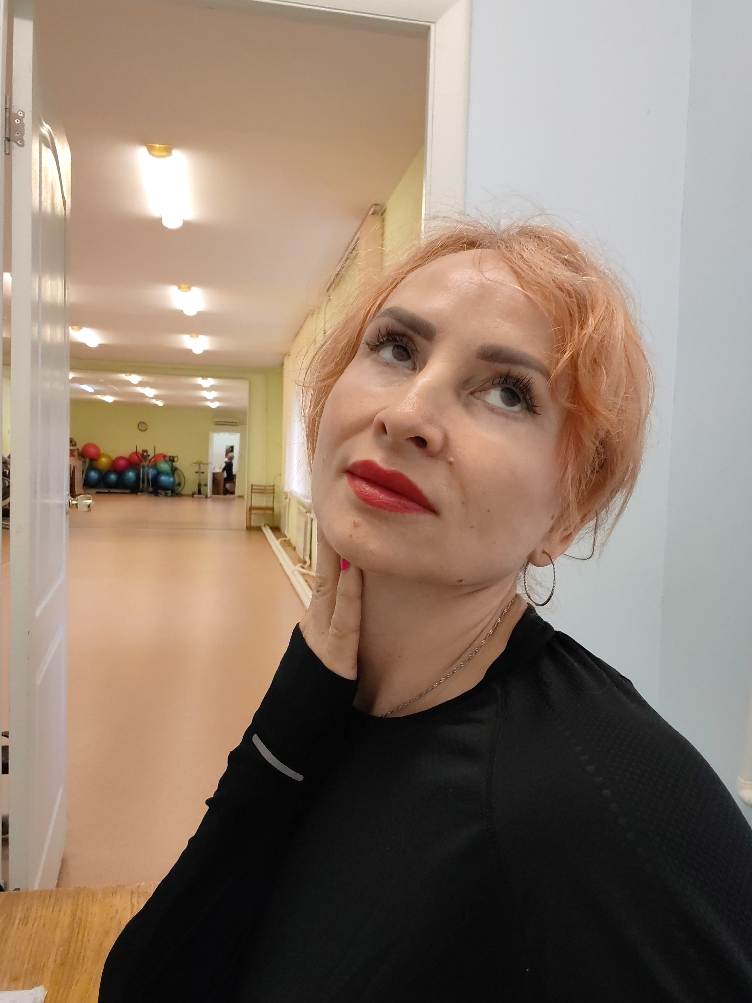 The image depicts a close-up of a woman with short, orange-dyed hair and red lipstick, looking upwards and to the right. She is wearing a black long-sleeved shirt and a silver necklace. Her right hand is touching her neck. In the background, a brightly lit room, possibly a gym or dance studio, is visible through an open doorway. This room contains exercise equipment, including stability balls and what appear to be stationary bikes. The environment suggests an indoor public space. The image was taken in Chaikovsky, Russia.