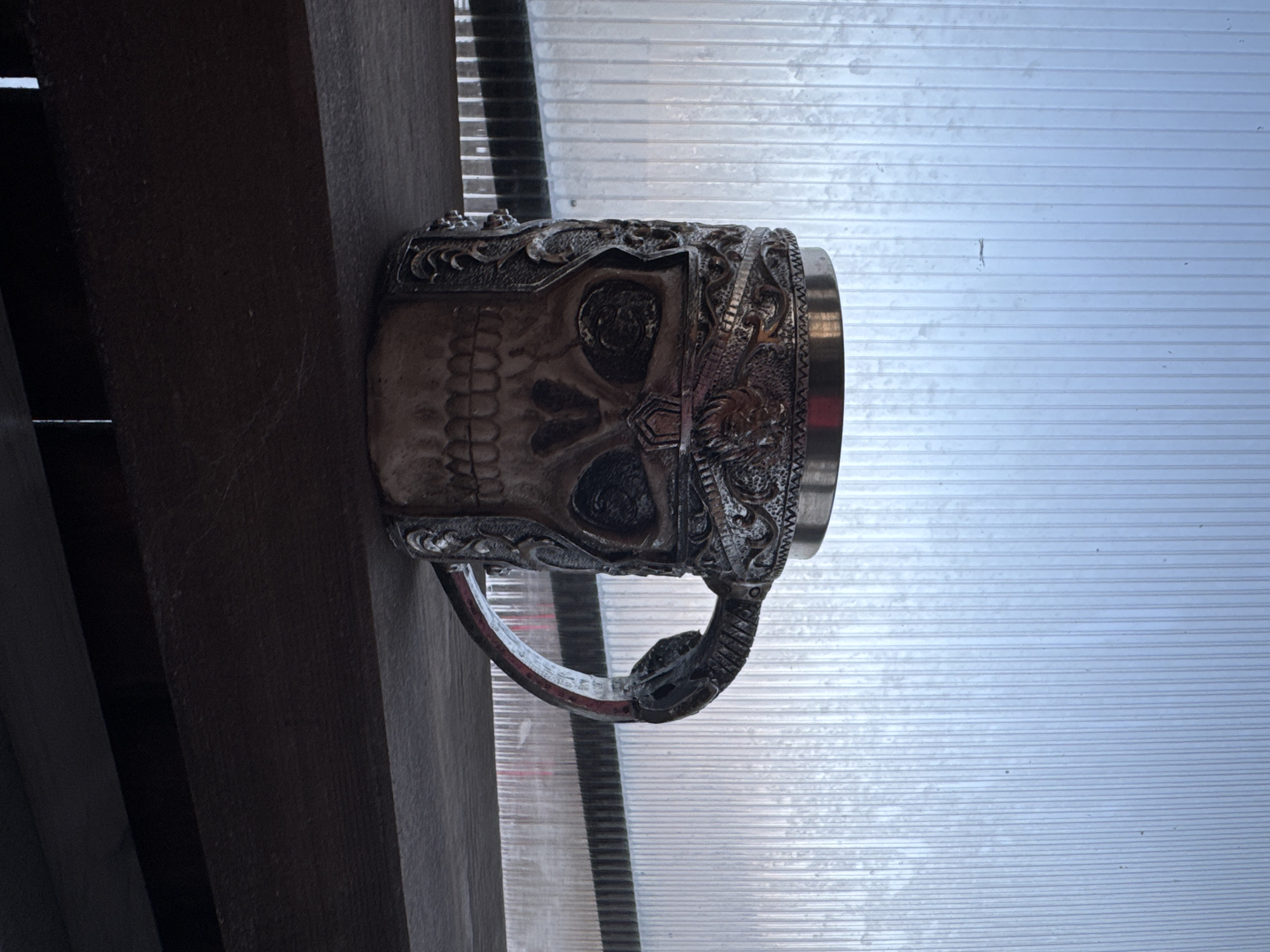 The image shows a detailed skull mug, resting on a wooden beam. The mug is primarily silver with intricate detailing, including a skull face and ornate patterns around the rim. The handle of the mug is shaped like a serpent. The mug appears to be outdoors under some type of awning, with corrugated plastic sheeting visible in the background. This scene is located in the city of Chaikovsky, Russia.