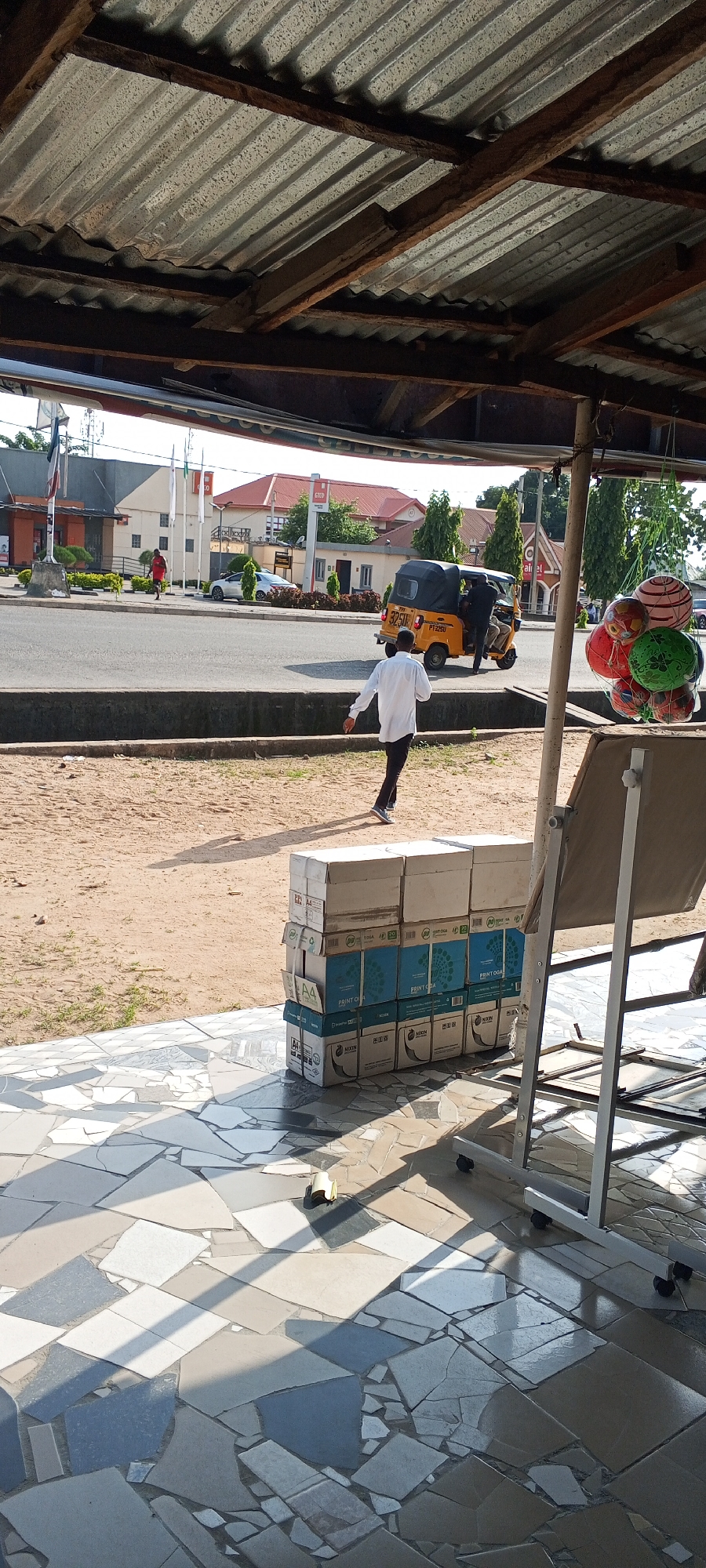 The image depicts a street scene in Jalingo, Nigeria, viewed from beneath a corrugated metal roof supported by wooden beams. In the foreground, a stack of cardboard boxes is piled on a tiled floor. The boxes are of various sizes and colors, some displaying printed text, including "A4" and "PRINT". Sunlight casts distinct shadows across the patterned tiles.
Further back, a man in a white shirt and dark trousers walks away from the camera towards a yellow auto-rickshaw, also known as a "Keke Napep", which is parked on the road. Another person is visible inside the auto-rickshaw. Across the street, a white car is parked near a building with an orange facade. Several other buildings, some with red roofs, and trees are visible in the background. Flags are also present, mounted on poles in front of some of the buildings. To the right of the image, a collection of colorful, spherical objects, possibly balloons or decorative items, are hanging from a string. The overall scene suggests a commercial or roadside area in a Nigerian city.