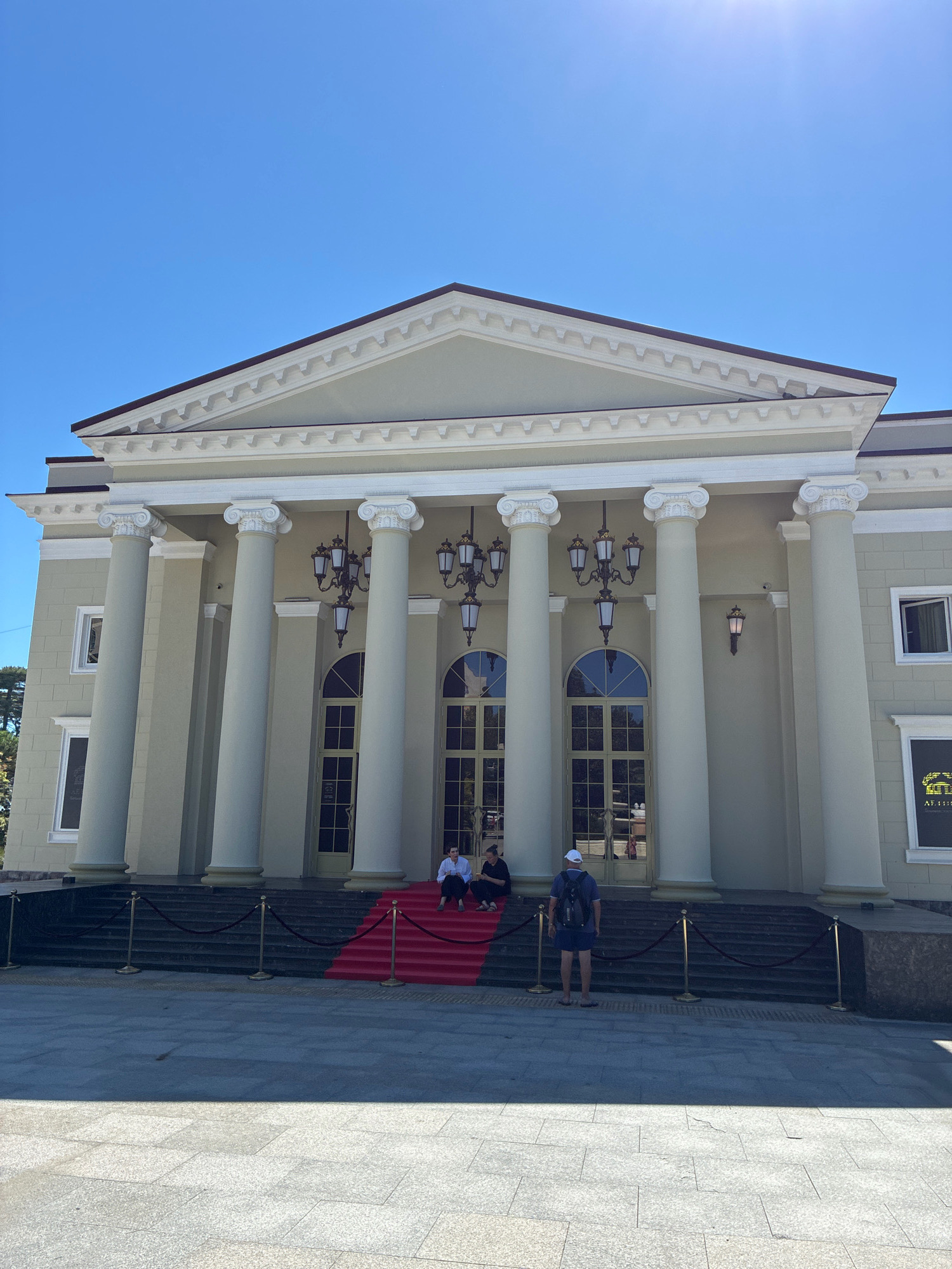 The image shows a grand building with large white columns and a triangular pediment under a clear blue sky. A red carpet leads up a set of stairs to the entrance. Several people are visible on the steps and in front of the building. The building appears to be the Sochi Sea Terminal, a prominent landmark in Sochi, Russia. The facade of the building is painted a light cream color, and decorative lamps hang from the ceiling between the columns. The ground in front of the building is paved with large square tiles. The overall scene suggests a formal or ceremonial occasion, possibly with visitors arriving or departing.
