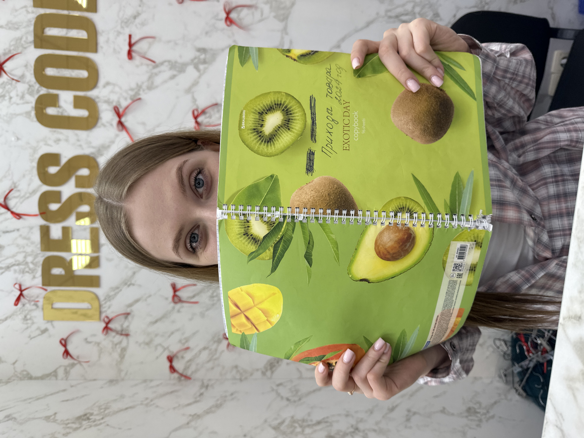 A young woman is depicted from the chest up, holding a green spiral-bound notebook with tropical fruit designs (kiwi, mango, papaya, avocado) in front of the lower half of her face. Her blue eyes are visible above the notebook. She has light brown hair and wears a plaid shirt over a white undershirt. Her hands, with light pink manicured fingernails, are visible holding the notebook; a ring is present on her left ring finger. The notebook cover displays the Russian text "Приходы товара 2024 год" (Goods Arrivals 2024 year), "EXOTIC DAY copybook," "96 sheets," and the "BRAUBERG" logo, along with a barcode and QR code.
The background consists of a white and gray marble-patterned wall featuring golden mirrored letters spelling "DRESS CODE" and numerous small red decorative bows. Clothing hangers are partially visible on the left side of the frame, and a dark office chair is partially visible on the right. A light-colored countertop or desk is in the foreground. The scene is located inside the "Dress Code" clothing store at Ulitsa Lenina, 61A, in Chaikovsky, Perm Krai, Russia.
