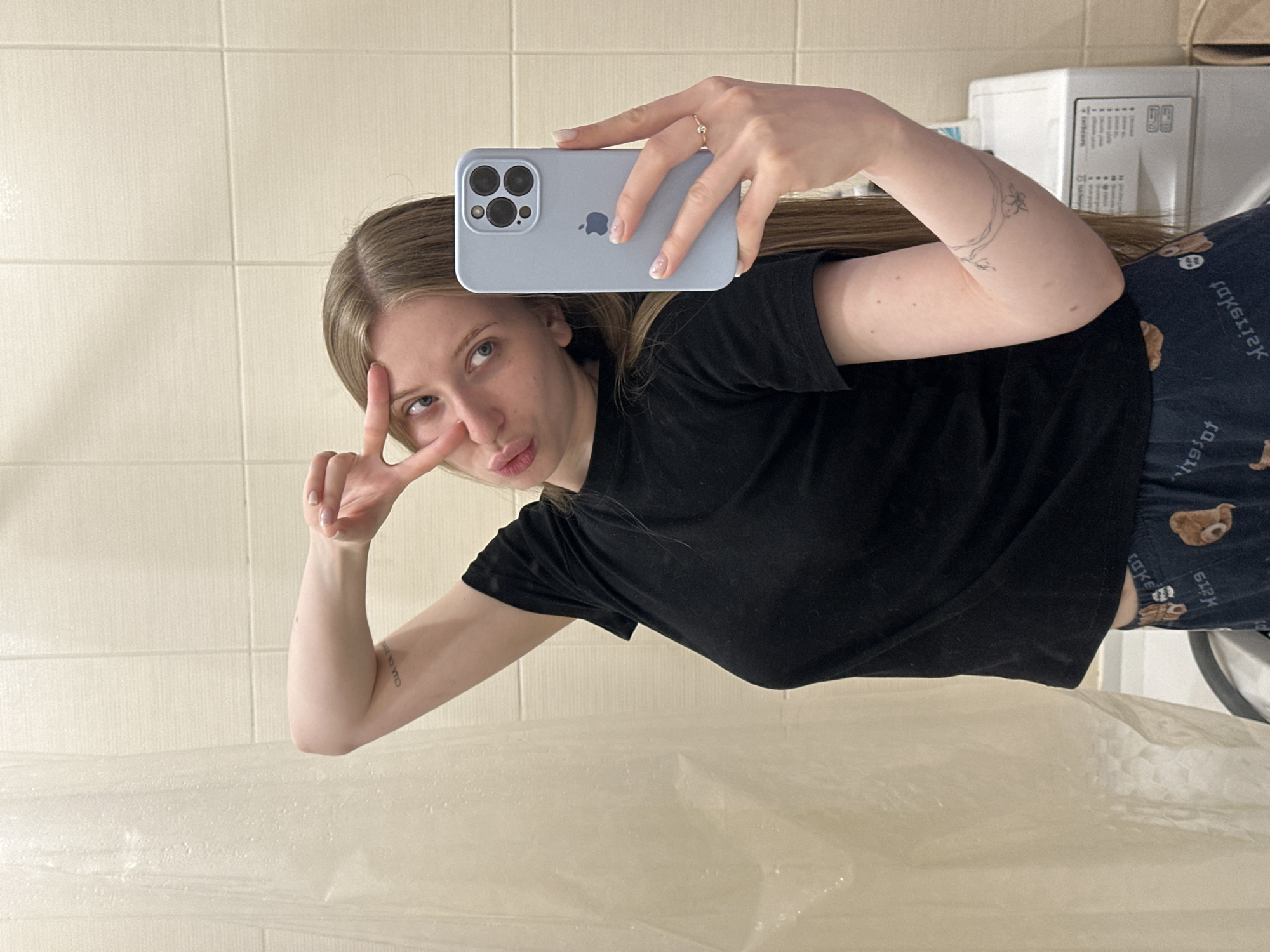 The image shows a young woman taking a selfie in a bathroom. The woman has long blonde hair and is wearing a black t-shirt and patterned pajama pants. She is holding a light blue iPhone 13 Pro, making a peace sign with one hand. The background features a tiled wall and a shower curtain. A microwave is visible in the background. The exact location within the city of Chaikovsky, Russia, cannot be determined from the image alone.