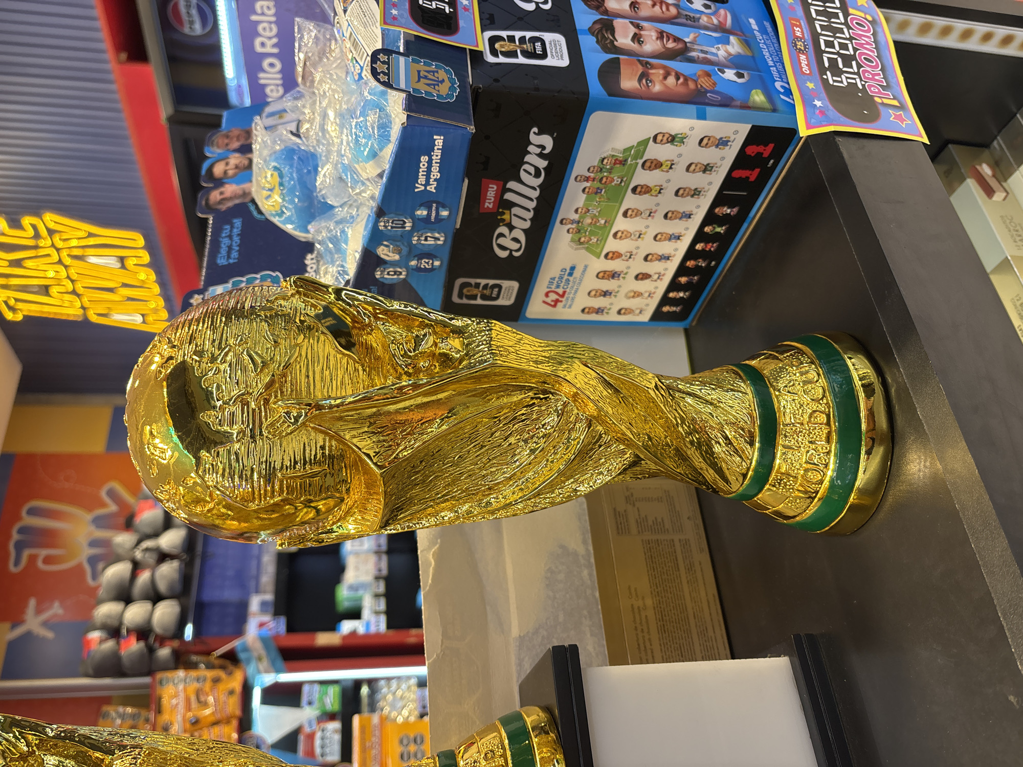A close-up, eye-level, indoor shot features a golden replica of the FIFA World Cup trophy prominently displayed on a dark, reflective surface. The trophy, with its signature globe and sculptural figures, gleams under bright lighting. To the right of the trophy, a stack of merchandise boxes is visible, including one labeled "ZURU Ballers" featuring miniature soccer player figures and portraits of famous athletes. Another box displays the Argentine flag and the words "Vamos Argentina!" Above these boxes, a neon "TAKE AWAY" sign is partially visible against a striped blue and red background. In the background, shelves are stocked with various products, including what appear to be bags of snacks and packaged goods. A small Argentine flag is also visible to the left of the trophy. The overall environment suggests a retail setting, possibly a convenience store or souvenir shop at Aeropuerto Internacional Ezeiza, Argentina, given the presence of the World Cup trophy and Argentine-themed merchandise. The lighting is bright and artificial, creating reflections on the trophy and the counter. No people are present in the immediate foreground or interacting with the objects.