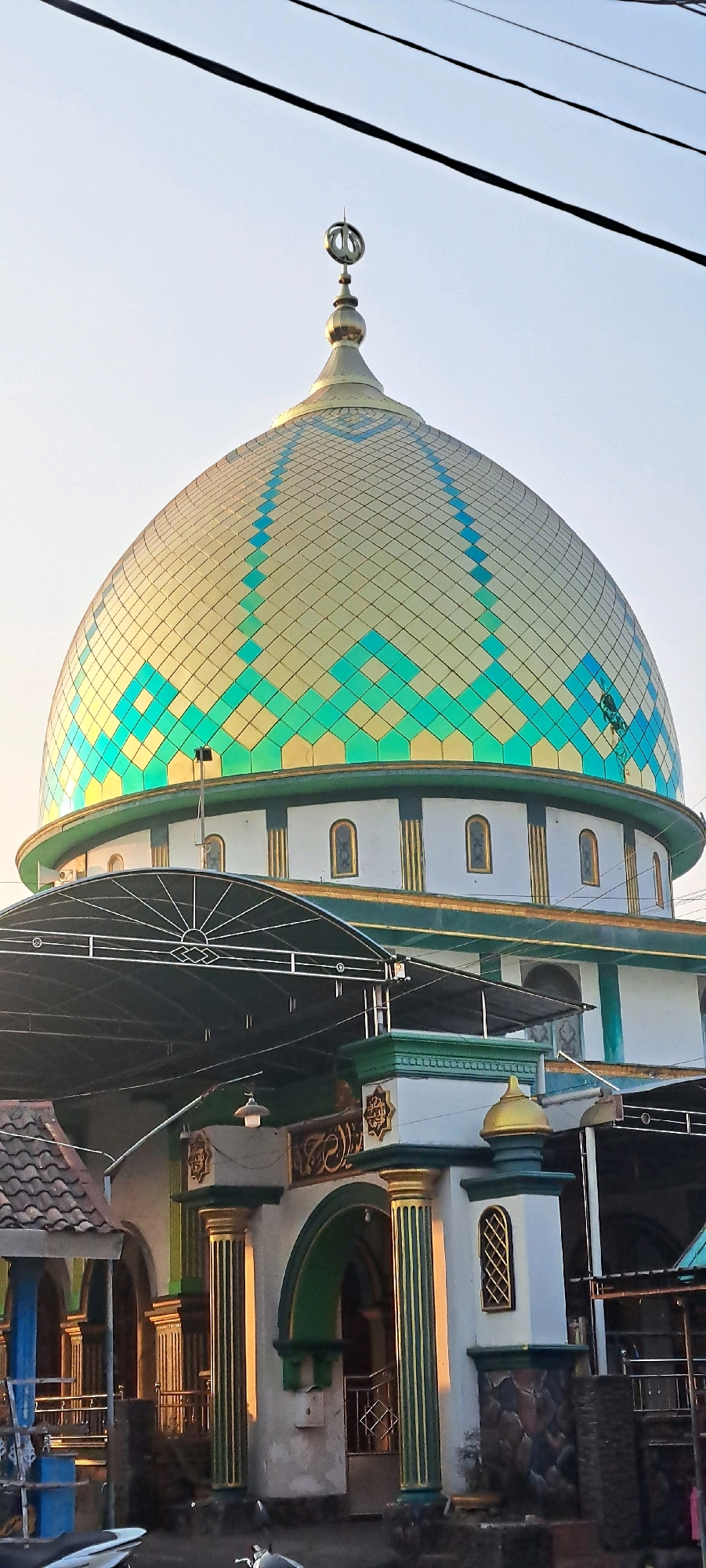 The image shows a mosque with a distinctive golden dome decorated with green and blue diamond patterns. The dome is topped with a finial. Below the dome, the mosque has white walls with green and gold decorative elements, including columns and arched windows. In the foreground, there is an entrance with large fluted columns and an arched gateway. A black awning covers part of the entrance area. The sky above is clear and light blue, with dark power lines running across the top of the frame. The overall impression is of a religious building, likely located in a populated area, given the presence of power lines and the urban-style architecture. The specific location mentioned, Kemiri Sewu, Indonesia, indicates a context within that country's architectural and cultural landscape.