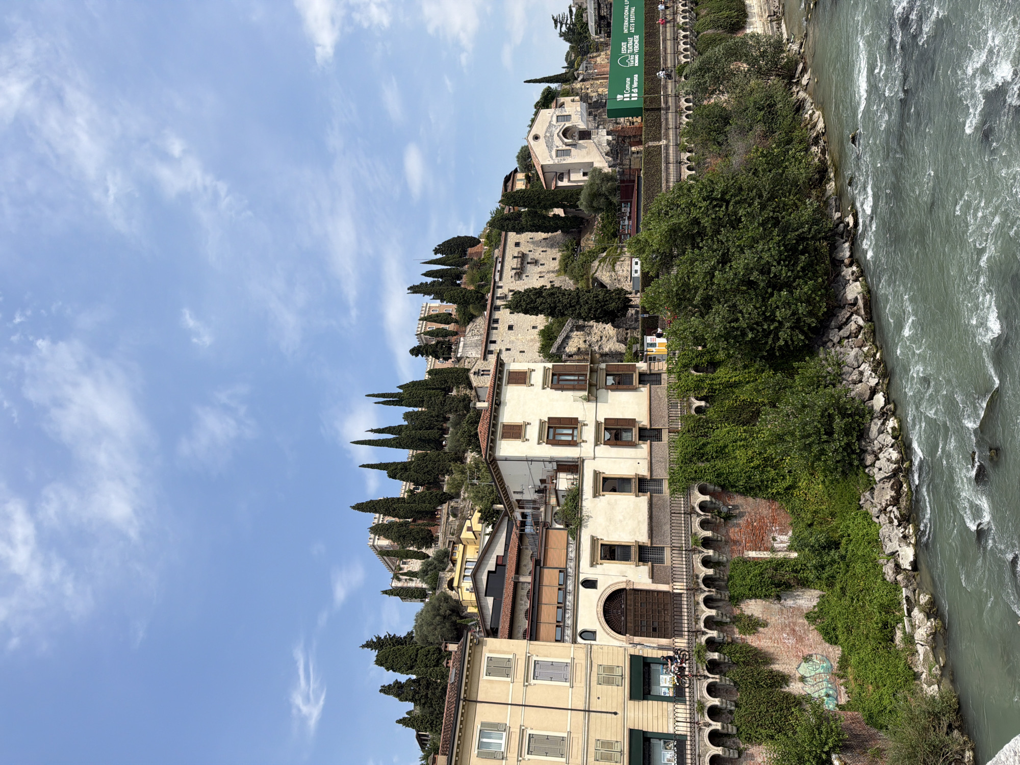 The image depicts a scenic view of Verona, Italy, featuring the Adige River in the foreground and a historic hillside cityscape on the opposite bank. The river flows with a moderate current, its greenish-gray water marked by white ripples and scattered dark rocks. Along the riverbank, there are large, rough stones and dense green foliage.
Above the riverbank, a stone wall with a series of arched openings runs horizontally across the midground, supporting a walkway adorned with railings. This wall is heavily draped in lush green ivy. Several multi-story buildings with light-colored facades and traditional tiled roofs are built into the hillside. Some of these buildings have prominent wooden shutters and decorative architectural details.
Cypress trees, tall and slender, are strategically planted along the slopes, interspersed with other greenery. Higher up on the hill, a more substantial stone structure, possibly a castle or fortress, is visible, with crenellated battlements and multiple levels. A small church with a domed roof and arched windows is also part of the elevated architecture.
A large green banner with text in Italian, including "ESTATE TEATRALE INTERNAZIONALE DI VERONA ARTS FESTIVAL," is displayed on the hillside near the buildings. In the lower left foreground, a single person is seen riding a scooter on the walkway near one of the buildings. Other individuals are subtly visible further along the walkway. The sky above is a clear, bright blue with scattered white clouds. The overall impression is one of historical grandeur and natural beauty.