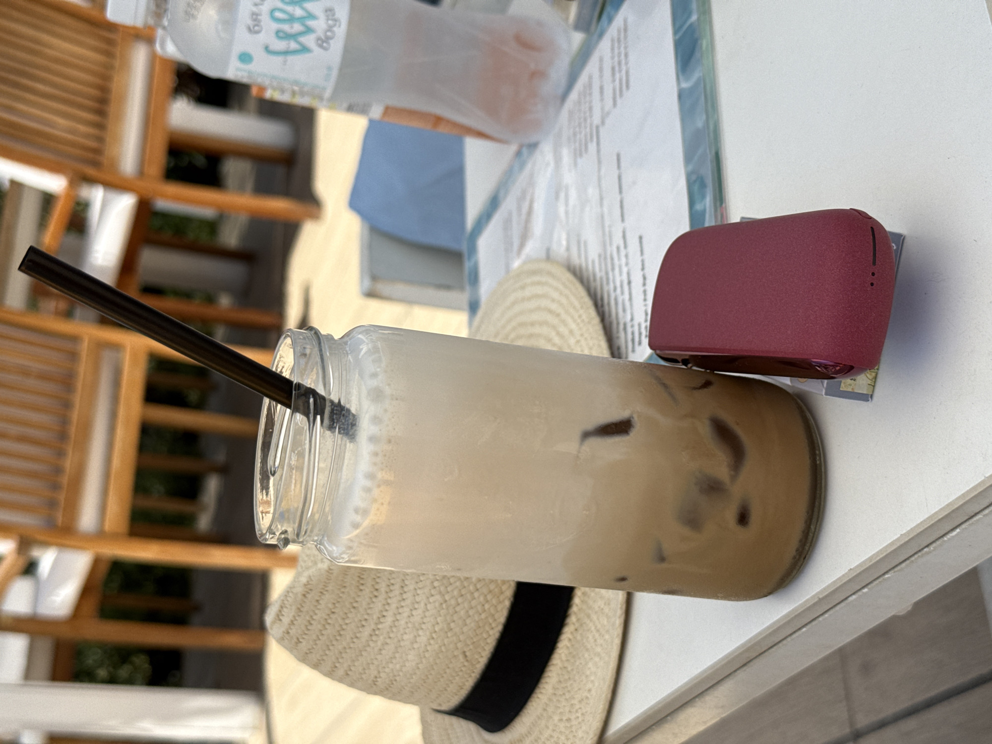 An iced coffee in a clear glass jar with a black straw sits on a white table, next to a straw hat with a black band. A red device with a matte finish is placed in front of the glass. In the background, wooden chairs with white cushions are visible, suggesting an outdoor seating area, possibly at a cafe or restaurant in Pleven, Bulgaria, given the context. A partially visible plastic bottle of water with a blue label and text, including the word "Boga," is also on the table. The scene appears to be captured in daylight, with a bright, natural setting.