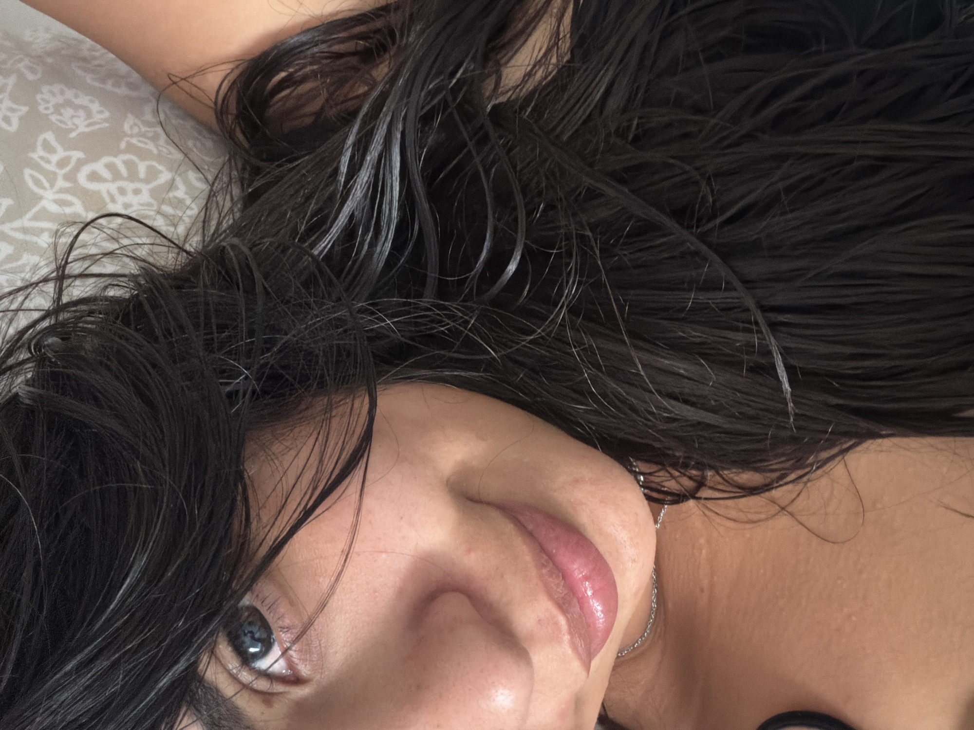 The image shows a close-up, low-angle selfie of a young woman lying down. She has long, dark, wet hair that frames her face. Her eye is visible, with blue irises and dark eyelashes. She has natural-looking skin with some freckles. Her lips are slightly parted and appear to have a glossy balm. She is wearing a delicate silver chain necklace. The background is a light-colored fabric with a subtle floral pattern, suggestive of bedding. The lighting is soft and natural, highlighting the texture of her hair and skin. There is no identifiable information within the image that would suggest a specific location in Duisburg, Germany, or any other city.