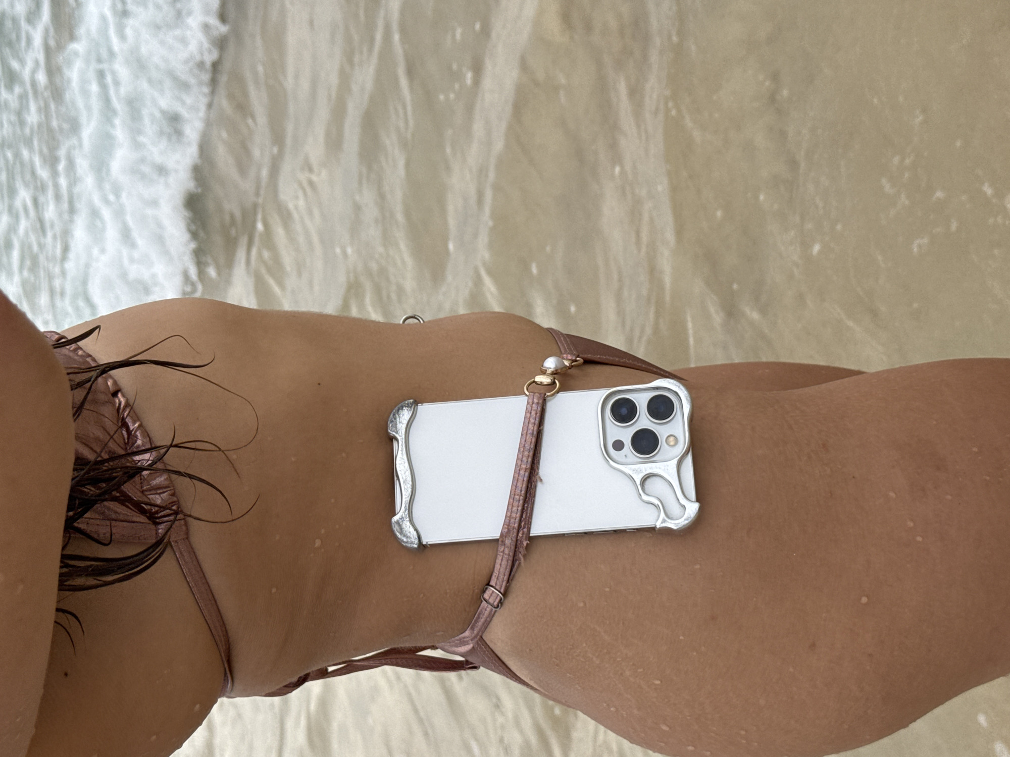 An individual wearing a rose gold bikini stands on a beach with gentle waves washing ashore. The person's side is visible, with a white smartphone attached to their bikini bottom strap. The phone is positioned on the person's hip, with the screen facing outward. The background consists of the ocean and the wet sand. This scene is located on the beach in Choeng Thale, Thailand.