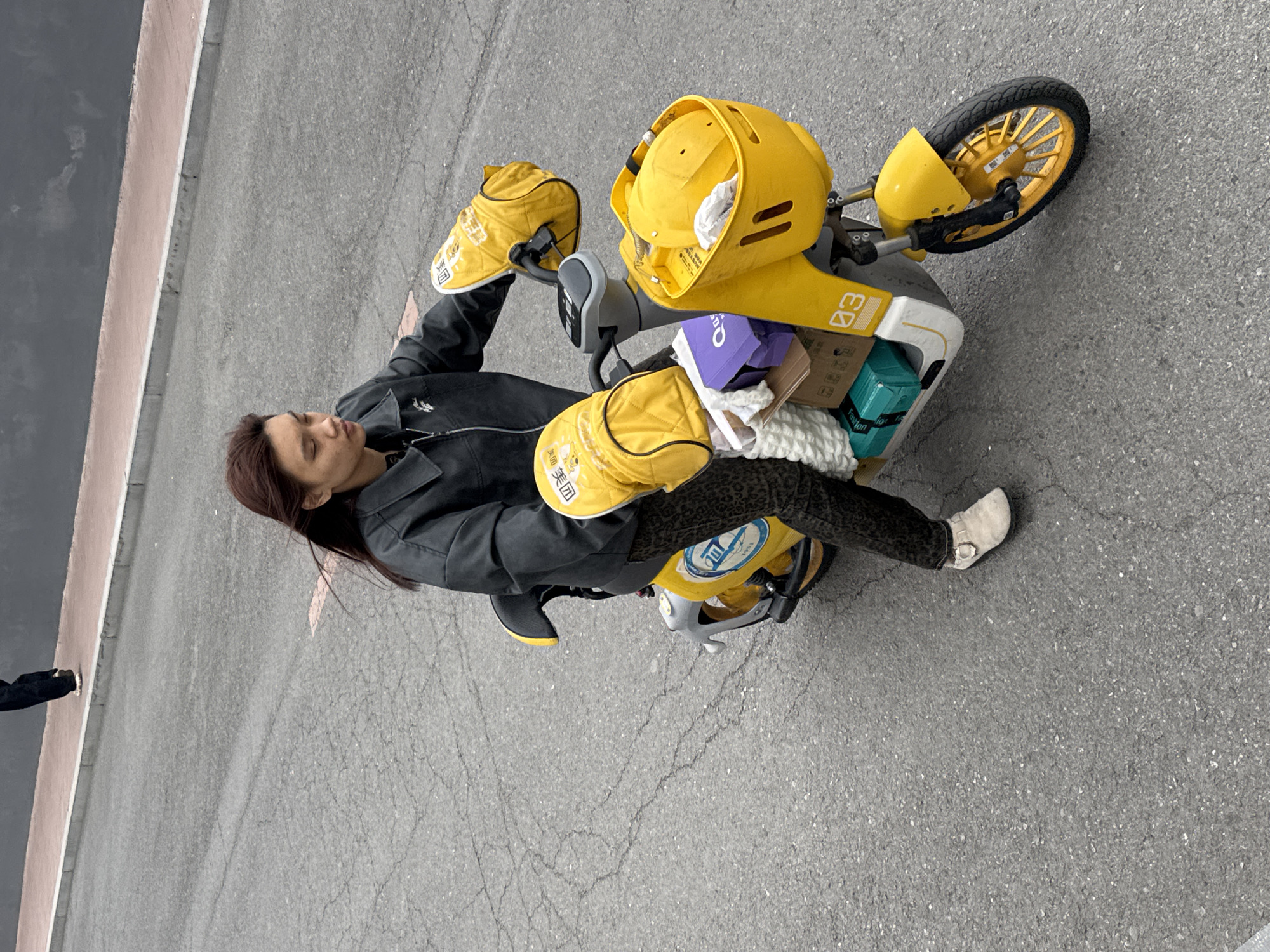 A woman is seated on a yellow electric scooter on a paved road. The scooter has a yellow helmet and yellow carrying bags attached to it, along with several packages strapped to the rear. The woman is wearing a black jacket, patterned leggings, and a white shoe. She is turned to her left, looking down at the scooter. The scooter appears to have the number "03" printed on its side. The location appears to be a street in Zhongtun, China.