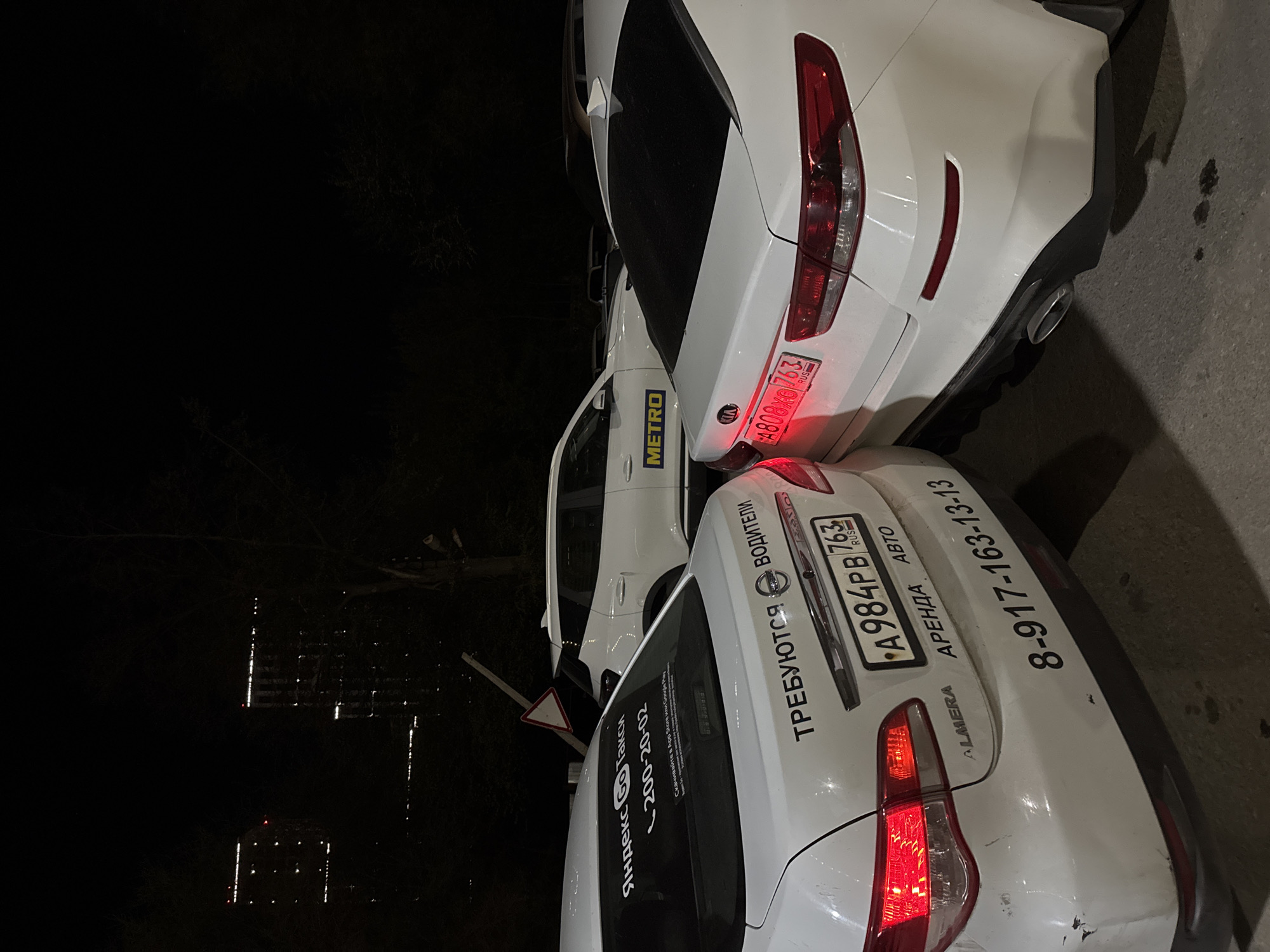 The image shows the rear of two parked white cars at night. The car on the right has the license plate "A984PB763", the word "ALMERA" near the taillight, and phone number "8-917-163-13-13". In Russian, the text on the car says "Drivers Required" and "Rent a Car". The car on the left has a "METRO" logo on the side and the license plate "A808OX763". In the background, there are buildings illuminated at night.