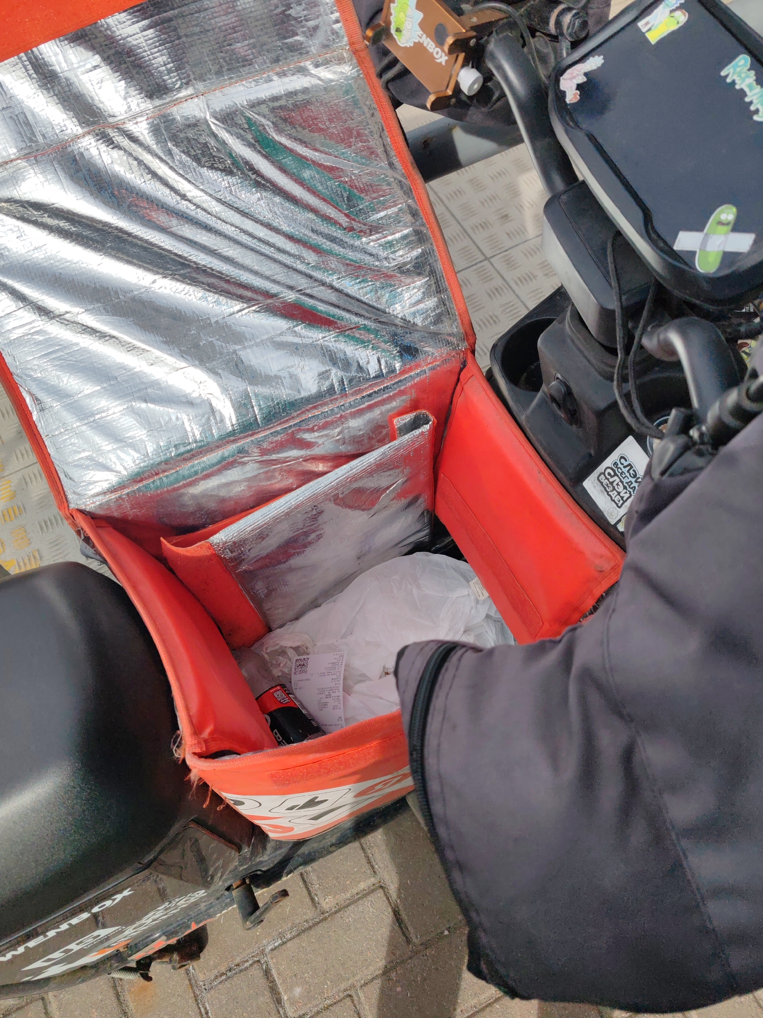 A delivery driver's open orange delivery bag on a scooter reveals a bottle, a white plastic bag, and a receipt. The scooter is parked on a brick sidewalk in Saint Petersburg, Russia, with a black-clad individual reaching into the bag.