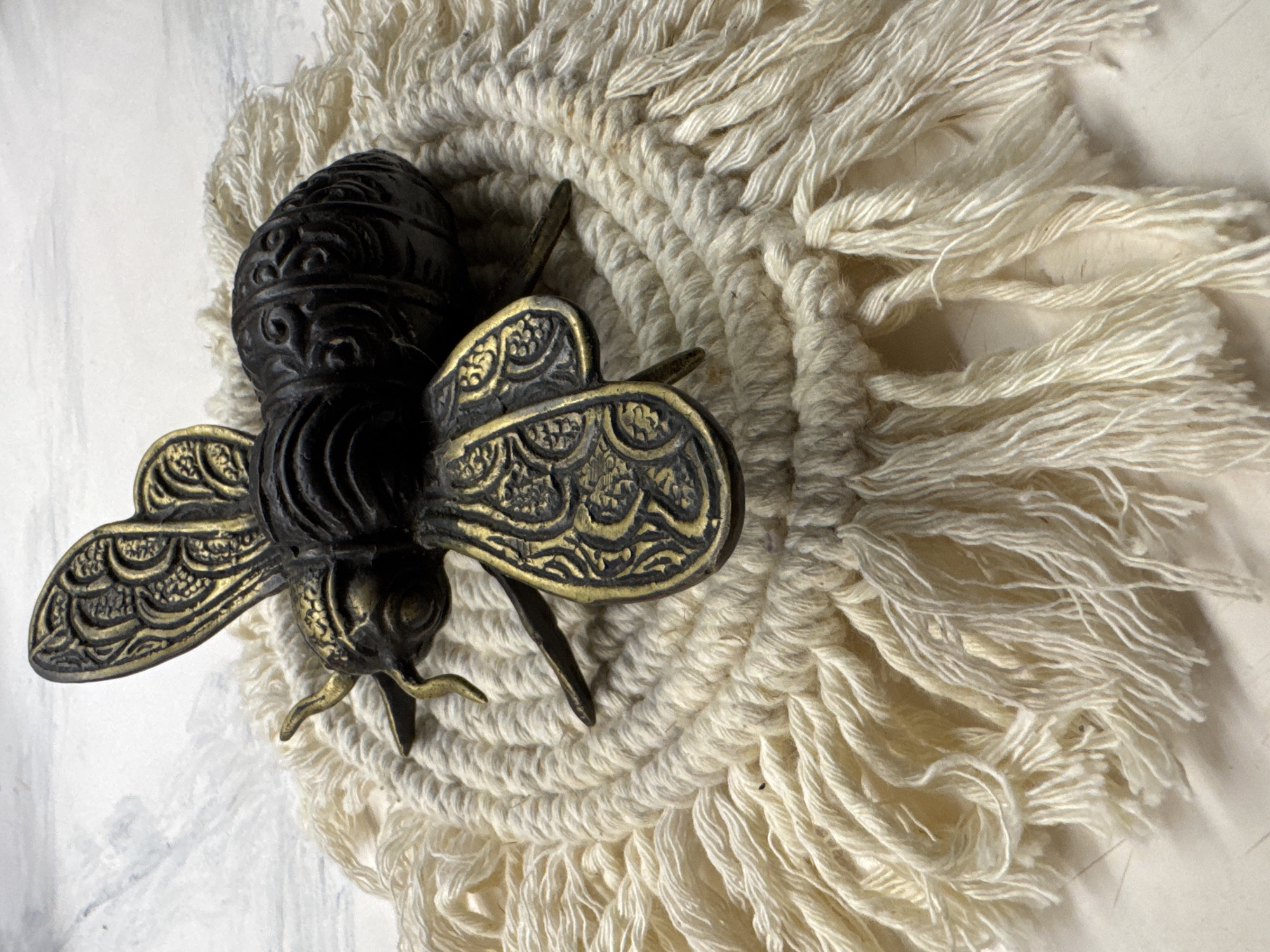 A high-angle, close-up shot depicts a metal bee figurine resting on a circular, fringed coaster. The bee is dark, possibly black, with gold-colored decorative wings. The coaster is made of a light-colored, possibly off-white, material with a thick, knotted center and long, loose fringe extending outward. The surface beneath the coaster appears to be a light-colored, possibly white, textured surface. The image does not include details that would confirm its location as Jimbaran, Indonesia.