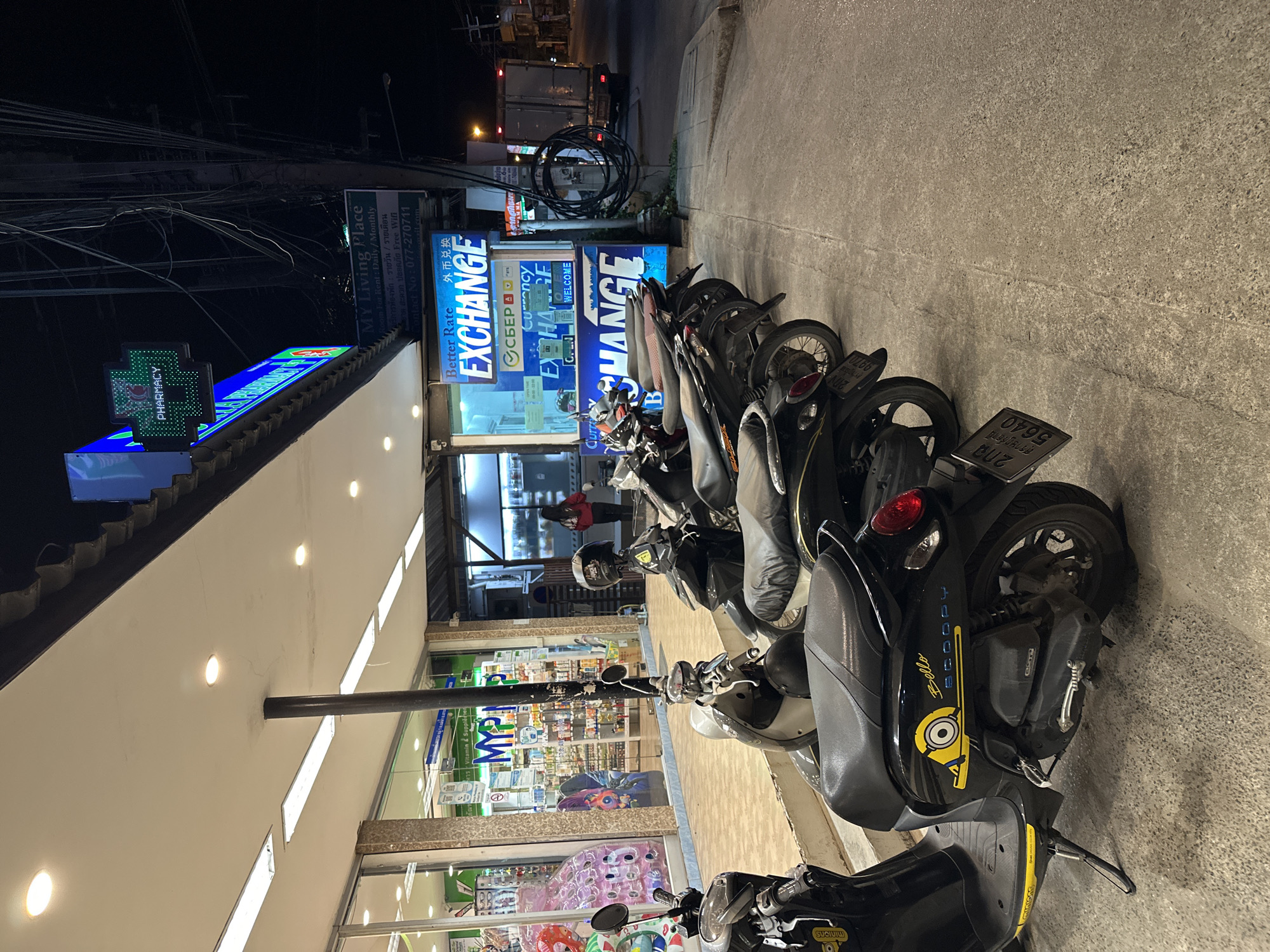 This is a photograph taken at night, likely on a street in Baan Mae Nam, Thailand, as indicated by the signage. The scene features a row of parked scooters lined up along a sidewalk in front of a storefront. The storefront appears to be a pharmacy, indicated by a glowing green cross-shaped sign with "PHARMACY" written on it, and a sign inside reading "MYP Vitamin & Supplements". Adjacent to the pharmacy is a currency exchange business with prominent blue and yellow signage that says "Better Rate EXCHANGE" in English, and also features Chinese characters for foreign currency exchange.
Several scooters are parked, with their license plates visible, some bearing Thai script. One scooter in the foreground has a Minion character sticker on it, along with the word "Bello" in yellow lettering. The lighting suggests it is nighttime, with streetlights and illuminated signs providing the primary illumination. The overall atmosphere is typical of a commercial area at night, with the scooters indicating a mode of transportation common in the region. No people are clearly visible in the foreground, though a person can be seen through the window of the pharmacy. The ground is paved concrete, and overhead wires are visible in the background, characteristic of urban or semi-urban environments.