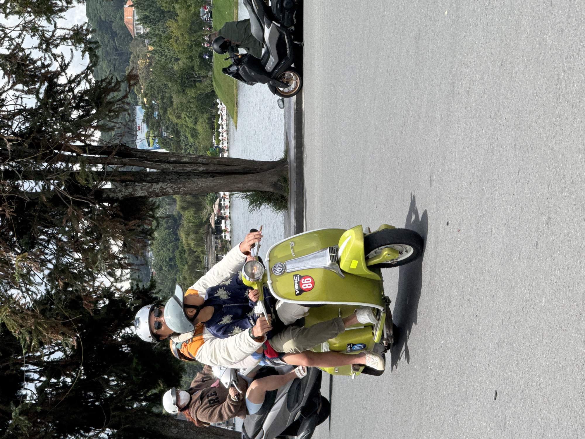 In Da Lat, Vietnam, on a bright day, three people ride two scooters along a paved road adjacent to a large body of water. The foreground features a bright green Vespa scooter carrying two individuals: an adult driver and a child passenger. The adult, wearing a white helmet and sunglasses, is dressed in a white jacket with orange accents. The child, also wearing a light blue helmet, is in the front, wearing a dark blue jacket with a star pattern. The scooter has a circular red decal with the number "86" on its side. Behind them, a second scooter, a dark grey motorcycle, carries a person with a white helmet and a face mask, wearing a brown hooded sweatshirt and light blue shorts. The backdrop reveals a picturesque scene with a calm lake, adorned with several white swan-shaped paddle boats. Lush green hills and trees line the far bank of the lake, with a few buildings visible among the foliage. The overall atmosphere is serene and leisurely.