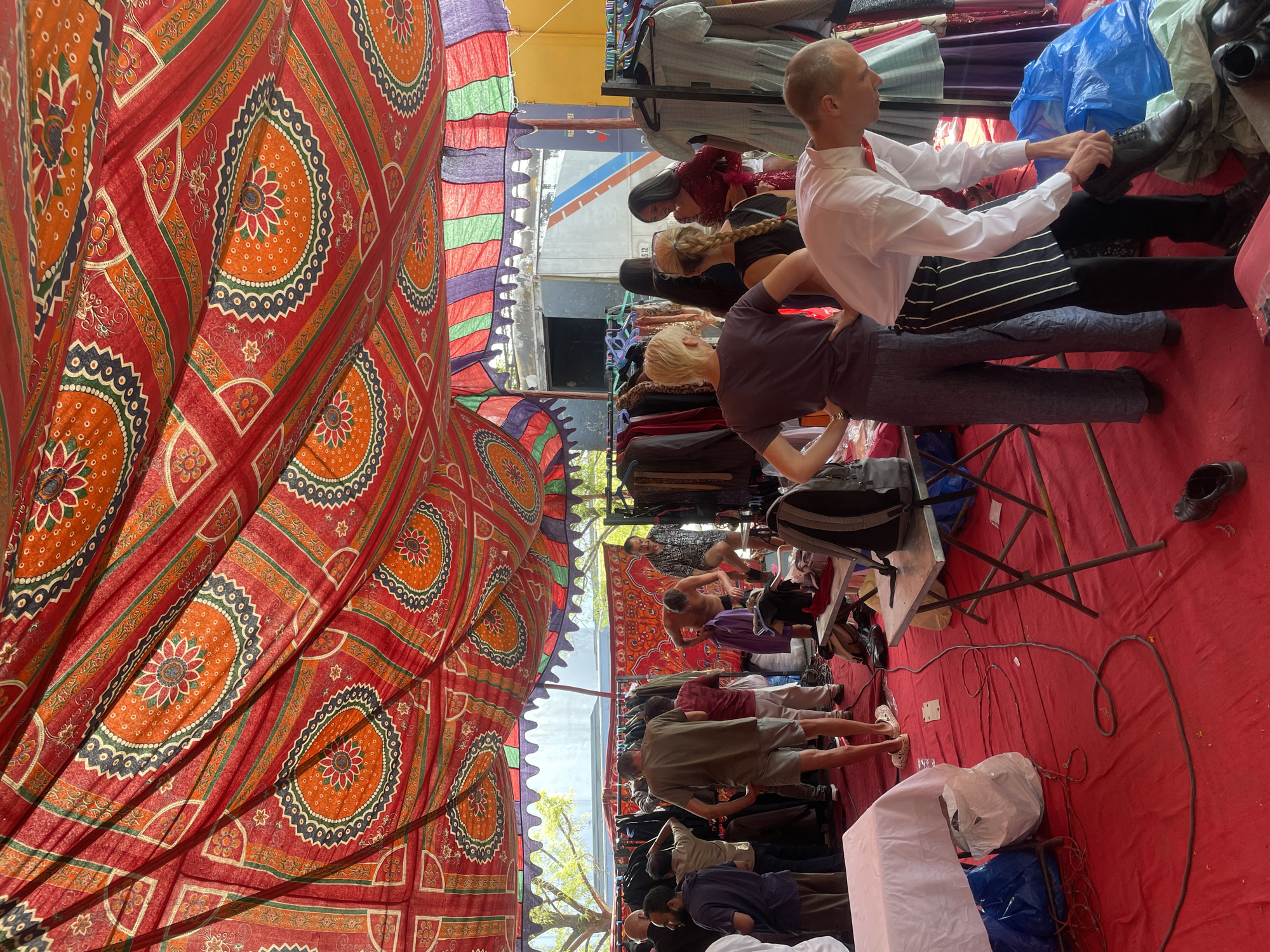 The image depicts the interior of a tent-like structure, possibly a marketplace in Anajpur, India, judging by the textiles. The tent is covered with red fabric featuring orange and blue floral patterns. Several people, dressed in varying attires, are browsing clothing hung on racks. One man, wearing a white shirt and a tie, is trying on a black shoe. Other individuals, including women with braided hair and men in casual clothing, are seen looking at the merchandise. The floor is covered in red fabric, and electrical cords snake across it. A rack and an electrical outlet are visible.