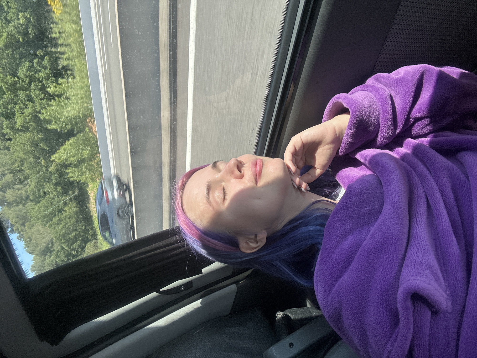 A person with purple and blue hair is asleep in a vehicle. They are covered with a purple blanket or robe. The window shows an outside view of a highway with vegetation on the side. The highway is likely in the vicinity of Pourcieux, France, given the user's specification. A car is visible on the road outside. The interior of the vehicle is visible, including the seat and window frame.