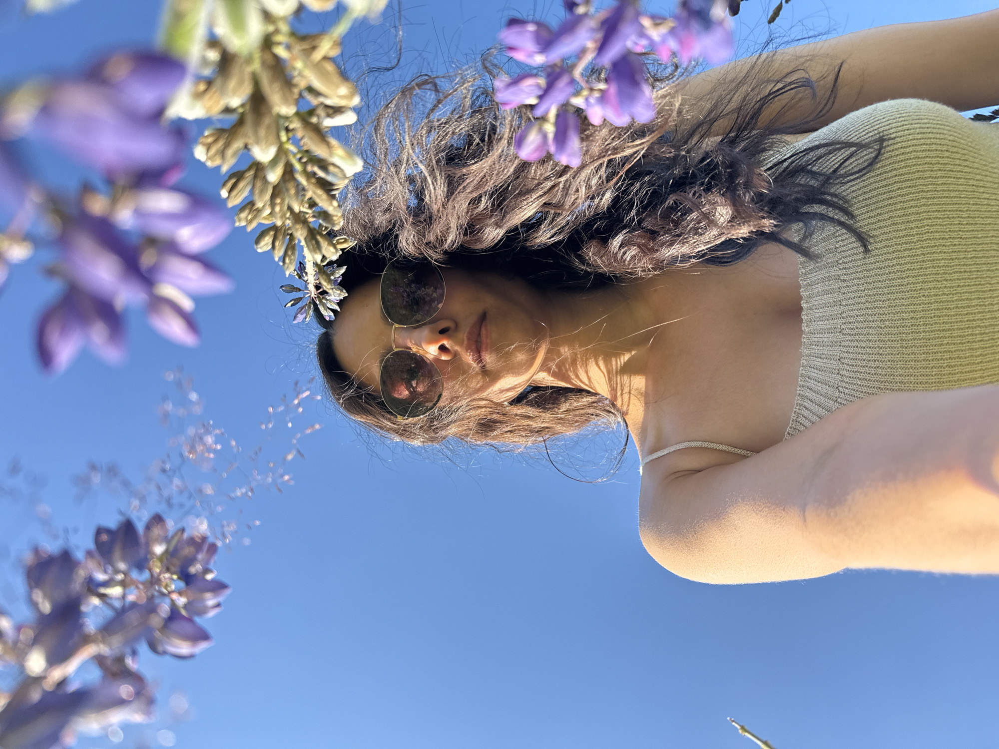 A low-angle shot shows a woman with long, dark, wavy hair and sunglasses looking upwards against a bright blue sky. She is wearing a light-colored, ribbed tank top. Purple flowers are in the foreground and background, some blurred and out of focus. The image appears to be taken in an outdoor setting, likely a park or garden, under natural sunlight. The presence of purple flowers suggests a possible connection to spring or summer. However, there is no information available in the image to determine the specific location within Chaikovsky, Russia.