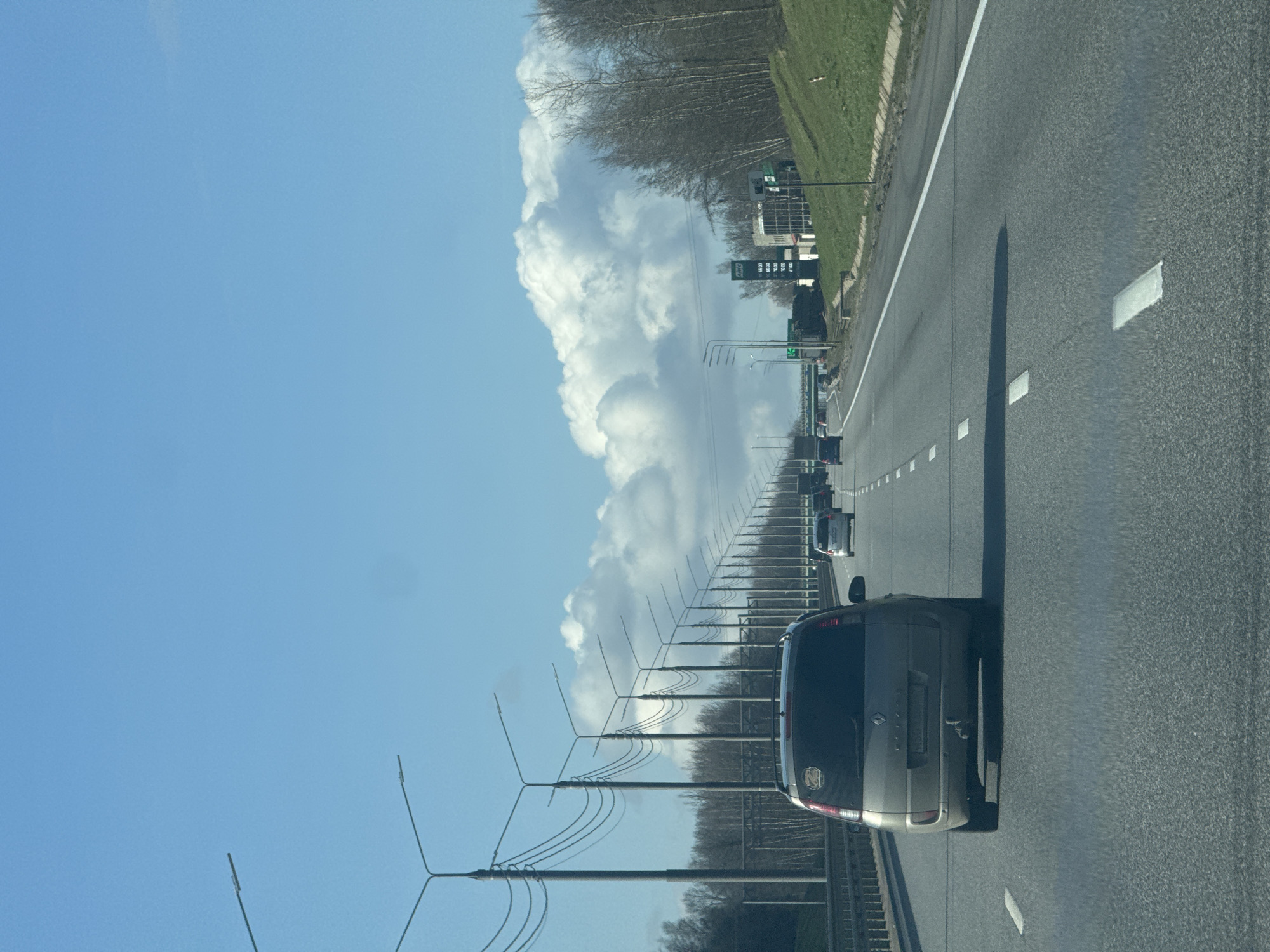 A landscape photograph depicts a multi-lane highway under a clear blue sky with cumulus clouds. A gray minivan occupies the right-hand lane, followed by other vehicles in the distance. Parallel to the road, a series of utility poles extend towards the horizon, their wires visible against the sky and clouds. A grassy embankment and trees border the highway on the right. In the middle distance, a gas station sign can be identified. The orientation of the image is sideways, requiring rotation for correct viewing. There is no information available to confirm it is in the city of Nikol'skoe, Russia.